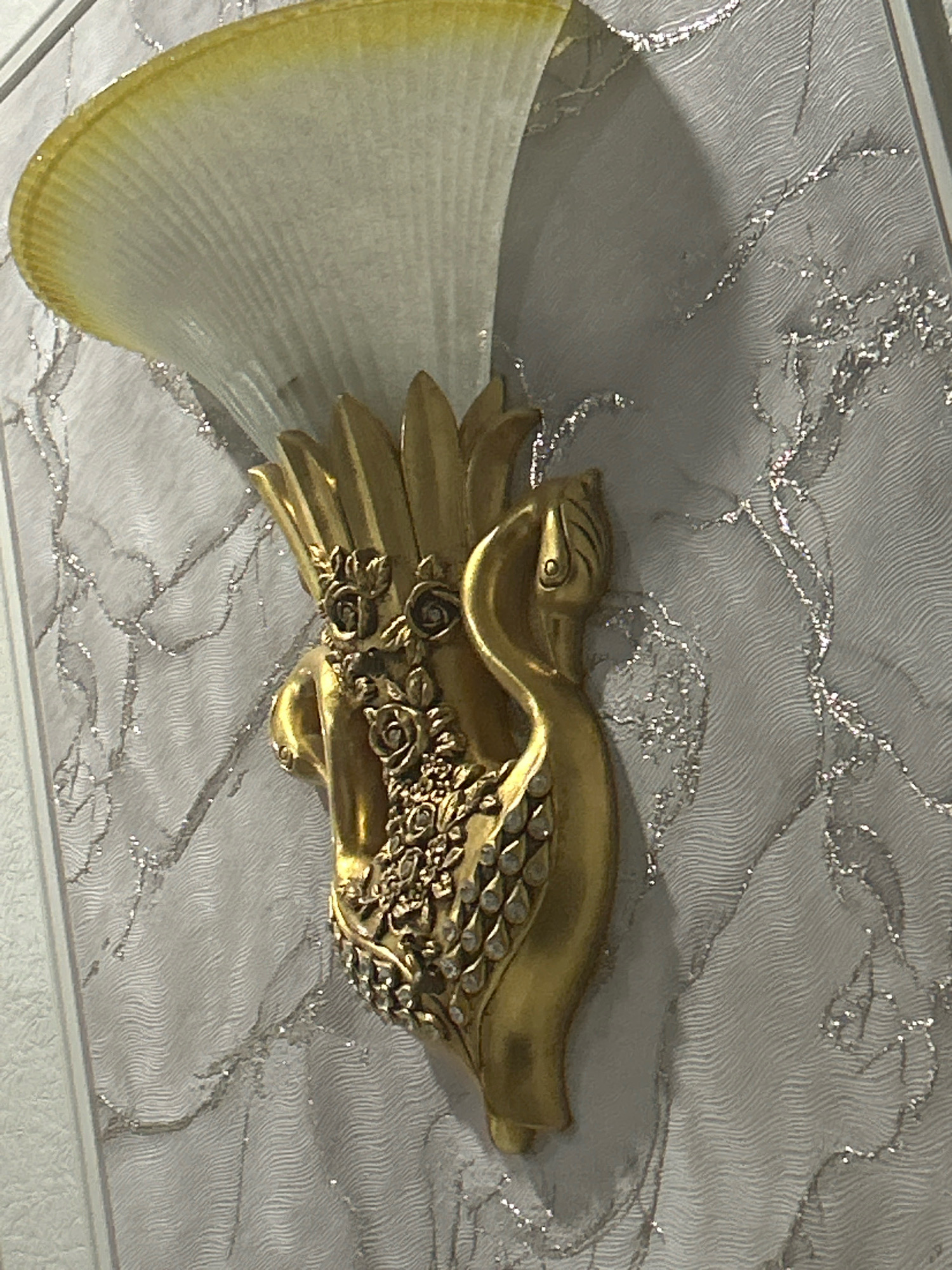 The image shows a close-up of a decorative wall sconce, likely made of gold-colored metal and frosted glass. The sconce is mounted on a wall with a textured, off-white wallpaper that has a subtle wave pattern and silver metallic accents. The sconce itself is ornate, featuring a stylized swan or bird motif with floral embellishments and what appear to be small, clear rhinestones. The glass part of the sconce is fan-shaped and appears to be illuminated from within, casting a soft yellow glow. The lighting suggests an indoor setting, possibly a home or hotel in Aktau, Kazakhstan, given the context provided. No people or actions are depicted.