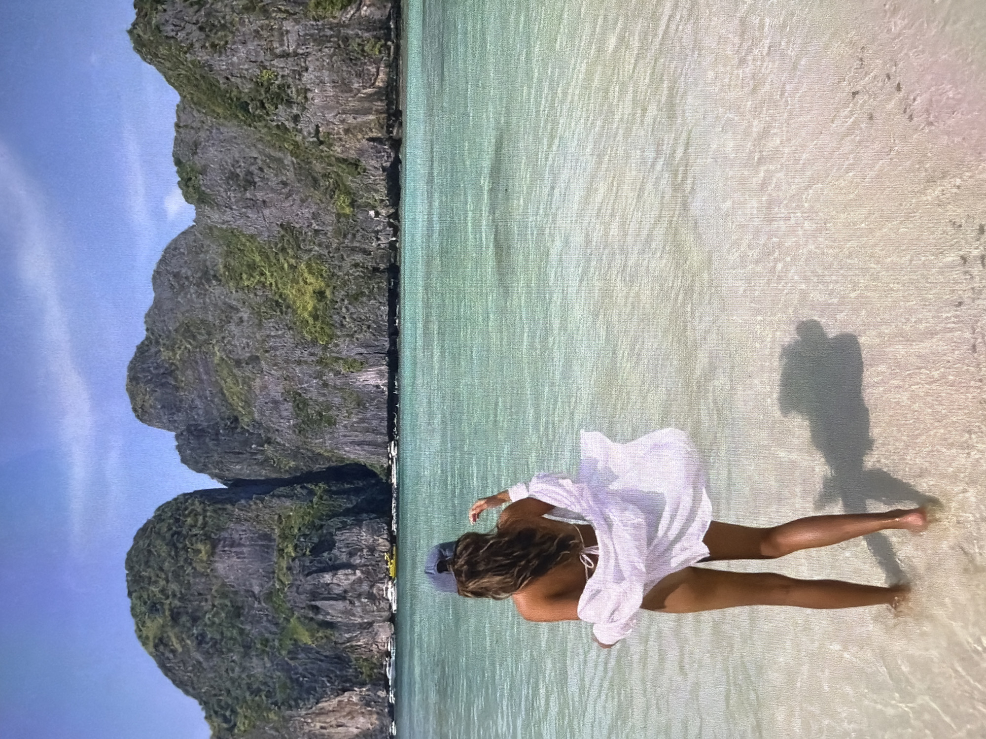 This image, presented sideways, captures a stunning tropical beach scene on a clear, sunny day. In the foreground, a person, viewed from behind, stands in very shallow, clear turquoise water, with their legs visible beneath the surface. They have long, wavy brown hair and are wearing a white, loose-fitting shirt or cover-up over a white bikini top, and a light-colored baseball cap. A dark shadow, likely cast by the photographer or another individual, is prominent on the sandy water in front of them.
The water extends into the mid-ground, showing gentle ripples and a pristine, light-colored sandy bottom. In the background, majestic, rugged limestone karsts or cliffs rise steeply from the sea, covered in patches of green vegetation, hinting at a lush environment. The sky above these formations is a bright, clear blue. In the far distance, at the base of the cliffs, several small boats can be faintly seen, adding to the sense of a popular yet serene destination. The overall scene conveys a feeling of relaxation and natural beauty in a tropical paradise. There is no visible text in the image.