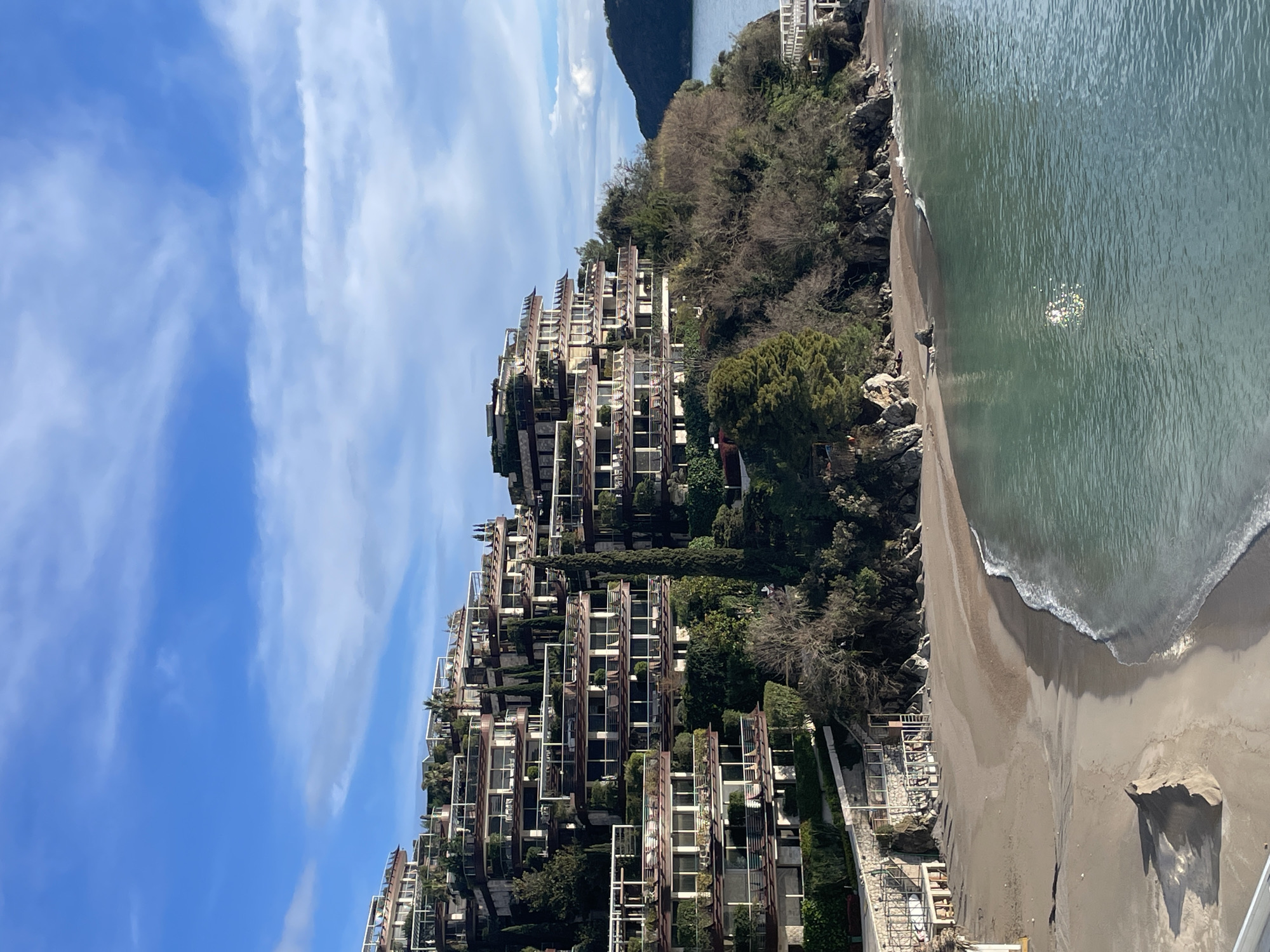 The image depicts a coastal scene in Budva, Montenegro, specifically showing the distinctive terraced architecture of the Sveti Stefan resort area. A large complex of multi-story buildings, characterized by a cascading, terraced design with numerous balconies and integrated natural vegetation, occupies a steep, forested hillside dominating the central and left portions of the frame. The hillside descends directly to a crescent-shaped sandy beach, devoid of human presence, located in the lower-left quadrant. The beach sand exhibits wave patterns and a distinct, conical sand accumulation in the foreground. The Adriatic Sea, calm and appearing in shades of light blue-green, occupies the right side of the image, with sunlight reflecting off its surface. Gentle waves break along the shoreline. A small white structure, possibly a pier or walkway, extends into the water from the base of the hill on the far right. The background features a distant, dark green landmass bordering the sea on the right. The sky is bright blue with scattered white, wispy clouds. No individuals or active subjects are visible within the scene.