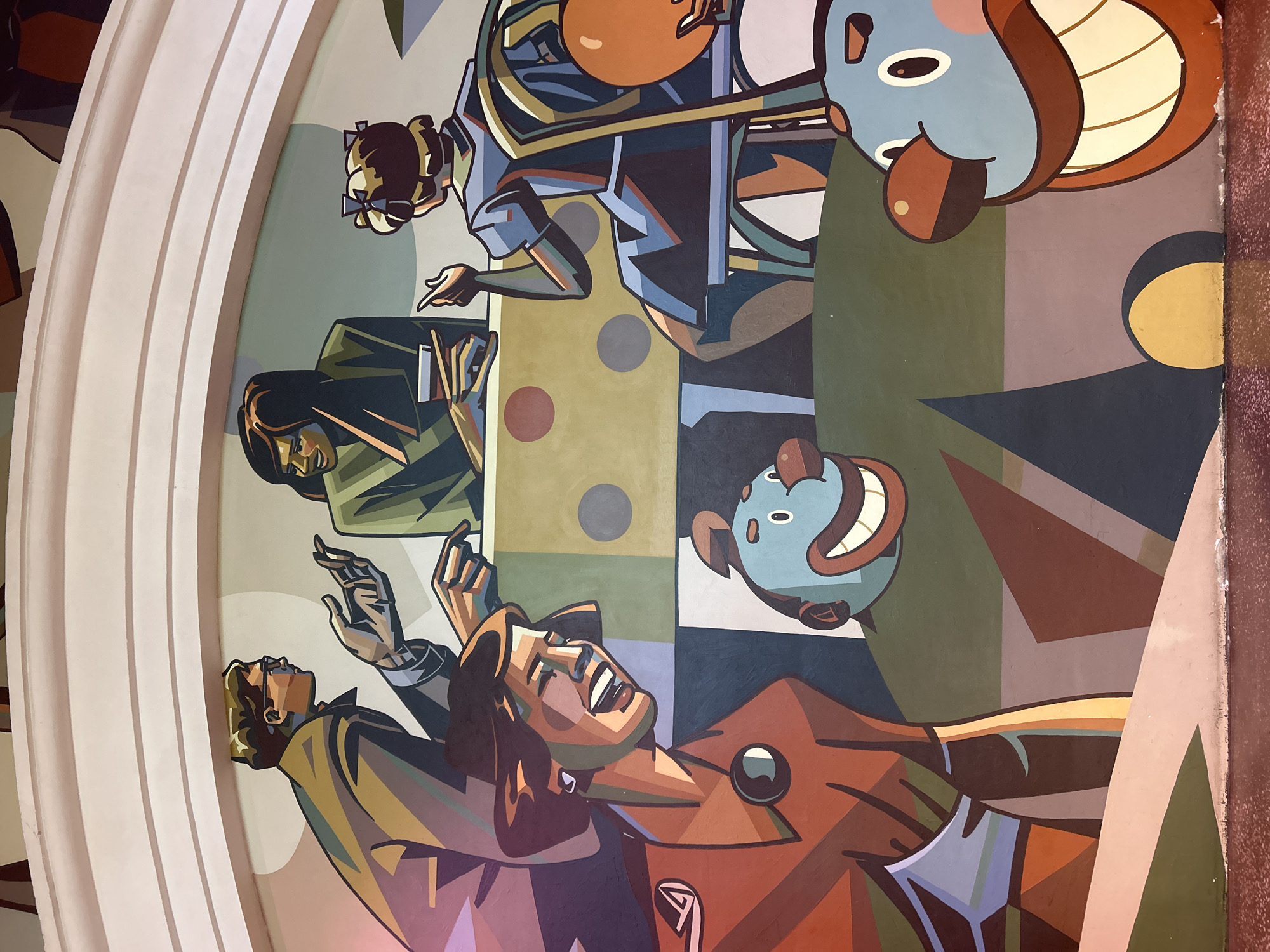 This is a mural painting depicting a group of people in what appears to be a social gathering or discussion. In the background, a man in a brown suit and glasses is gesturing with his hands, facing a woman in a green jacket who is holding a drink. To their right, a woman in a blue dress is seated and looking away. In the foreground, a woman in an orange-brown jacket is laughing heartily, looking towards the viewer. In the lower part of the image, two cartoonish clown-like characters with large smiles are visible, one to the left and one to the right, partially cropped. The background features geometric shapes and a patterned table. The style is characterized by bold lines and flat, colorful planes. The location is unknown.