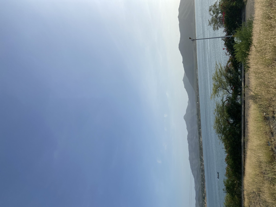 A serene landscape features a body of water stretching alongside hazy mountain silhouettes under a vast blue sky. The shoreline is lined with greenery, including trees, bushes with red flowers, and tall grass. A lone utility pole stands near the water's edge. A small boat floats on the calm water. The sky transitions from a deeper blue to a lighter shade near the horizon.