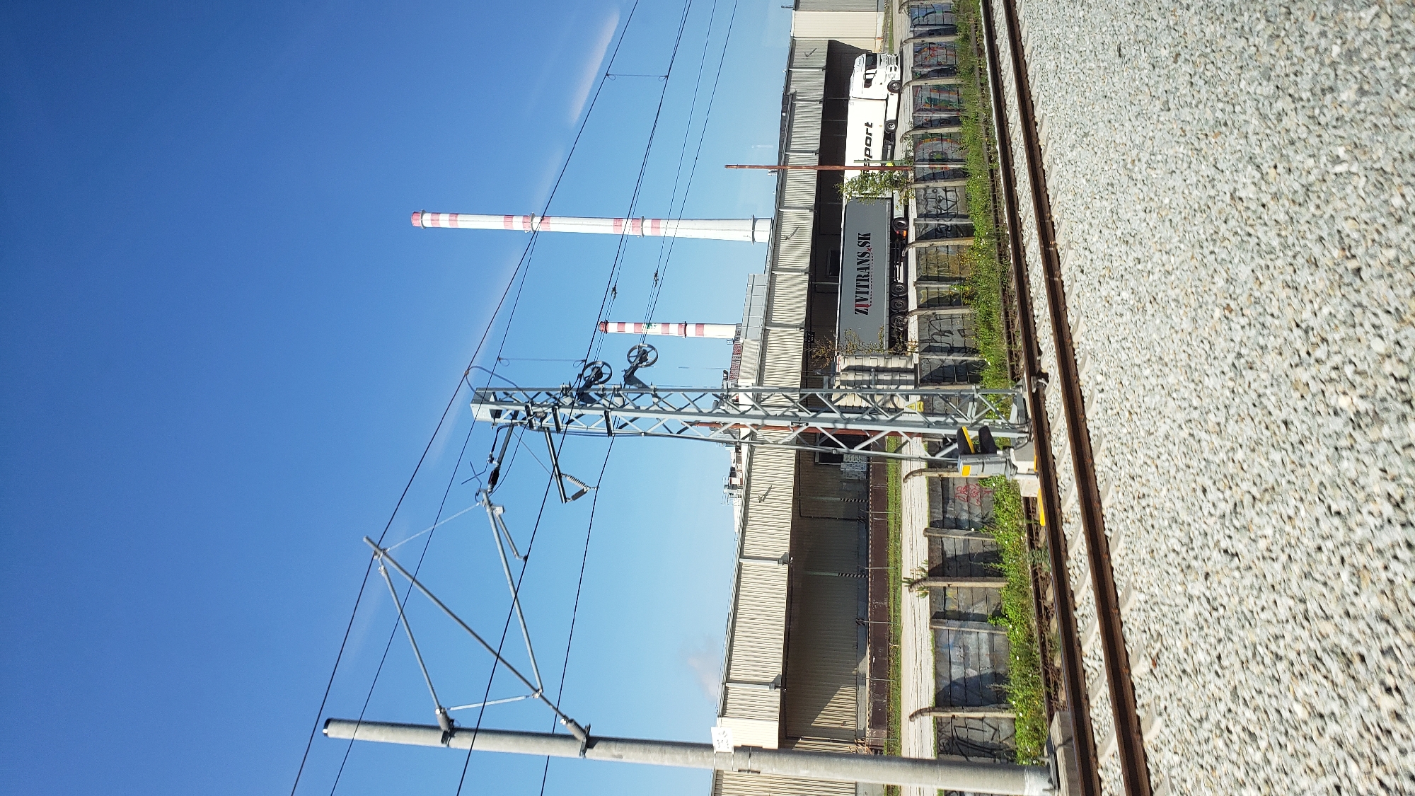 The image depicts an industrial area with clear blue skies and a bright sunny day. In the foreground, train tracks made of ballast are visible, running horizontally across the frame. A concrete pole with electrical lines for a train is situated to the left. Further back, a long industrial building with a light-colored, ribbed facade stretches across the middle ground. To the right of the building, two tall, red and white striped chimneys rise into the sky. In front of the industrial building, a gray truck with "ZIVITRANS.SK" written on its trailer is parked, alongside another white truck. A fence made of concrete blocks and barbed wire separates the foreground from the industrial area. The scene is set in Žilina, Slovakia.