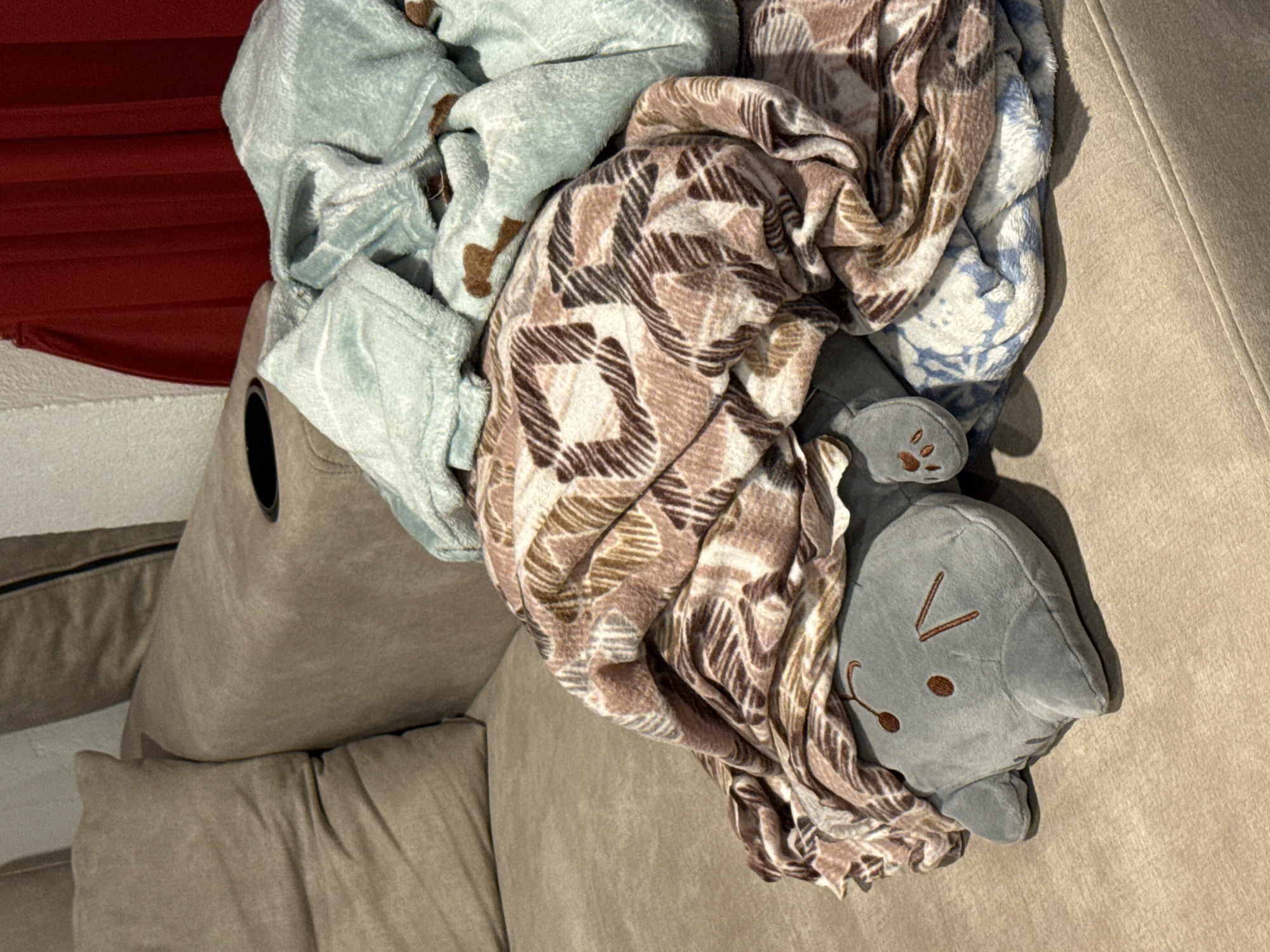 In Ko Samui District, Thailand, a plush gray cat toy is nestled on a beige sofa, partially covered by a patterned blanket and a lighter blue blanket. The cat toy has embroidered facial features and paws. A cup holder is visible on the armrest of the sofa. In the background, a portion of a red curtain is visible. The scene appears to be indoors, likely in a residential setting.