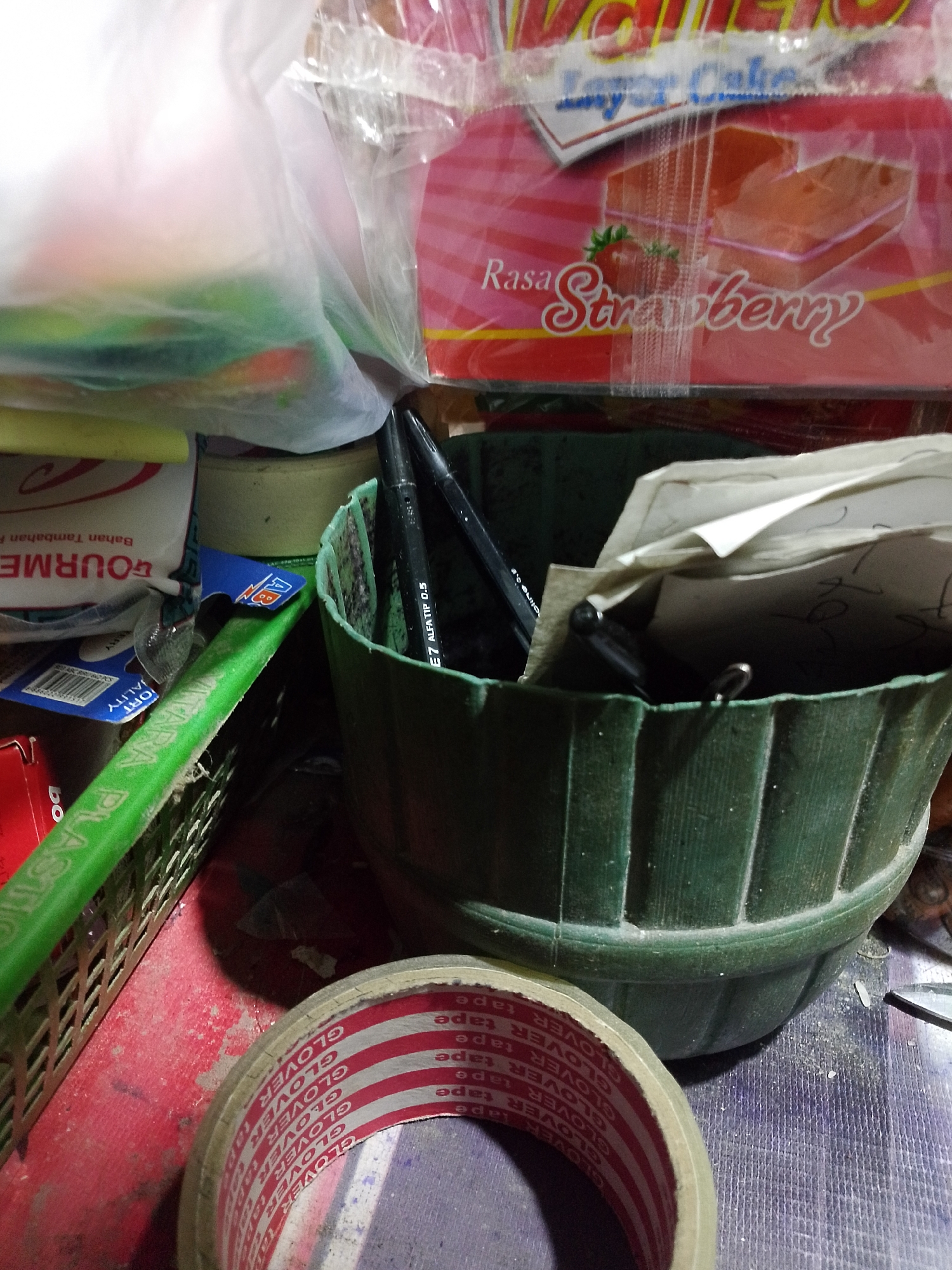 A close-up, low-angle, indoor shot depicts a collection of assorted items on a cluttered surface, possibly a workshop or storage area. In the foreground, a roll of packing tape with red and white striped detailing and the "GLOVER TAPE" logo is prominently featured. To the left, a bright green plastic handle of an unseen object rests on a woven basket. Behind this, a white paper bag with red text, possibly a food product labeled "GOURMET," is visible.
Further back, a green plastic pot contains several items. Two black pens with white markings are sticking out of the pot. One pen displays "AE7 ALFA TIP 0.5." Within the pot, a stack of papers, some with handwritten notes and numbers, is partially visible. A pink and red-patterned box, likely for food items, is visible at the top of the frame, with text that appears to read "Rasa Strawberry." The overall environment suggests a casual, perhaps domestic or small business setting. The lighting is dim, casting shadows and highlighting the textures of the objects. There are no people or clear indications of the specific location within Palembang, Indonesia, beyond the potential origin of the products.