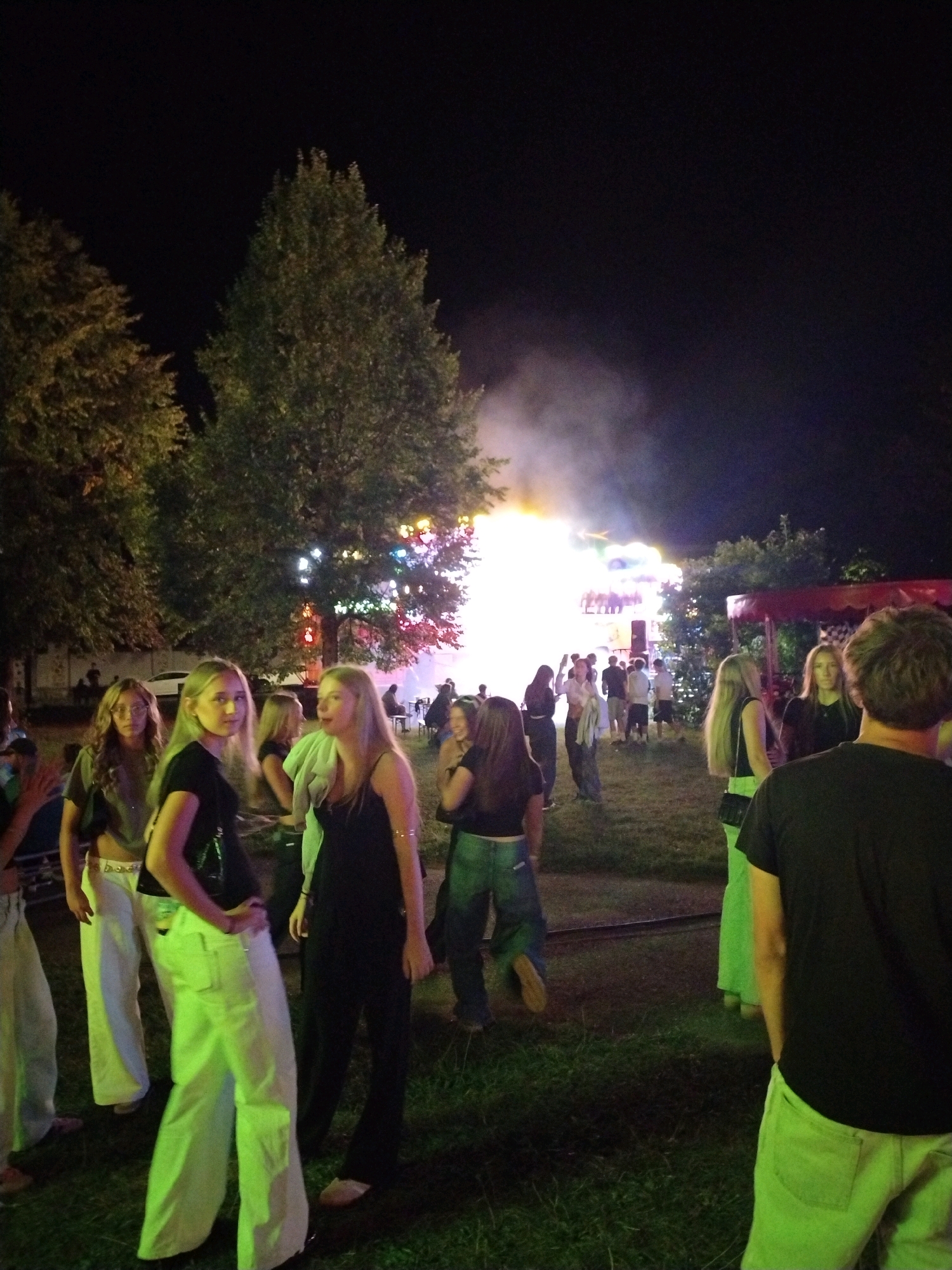 In the city of Písek, Czechia, a nighttime outdoor event is underway, likely a fair or festival, as indicated by the presence of illuminated structures and a crowd of people. In the foreground, four young women are visible. The woman on the left is wearing a dark top and light-colored, loose-fitting pants. Next to her, a woman in a black top and similar light-colored pants poses with her hand on her hip. Further to the right, another woman in a black top is partially obscured. The fourth woman, in the far right foreground, has her back to the camera, wearing a black t-shirt and light-colored pants. In the background, the event appears to be in full swing, with numerous people mingling and a bright, hazy light emanating from what seems to be a stage or attraction. Trees with colorful lights strung between them suggest a festive atmosphere. The ground is covered in grass, and the overall lighting is dim, characteristic of an evening event.
