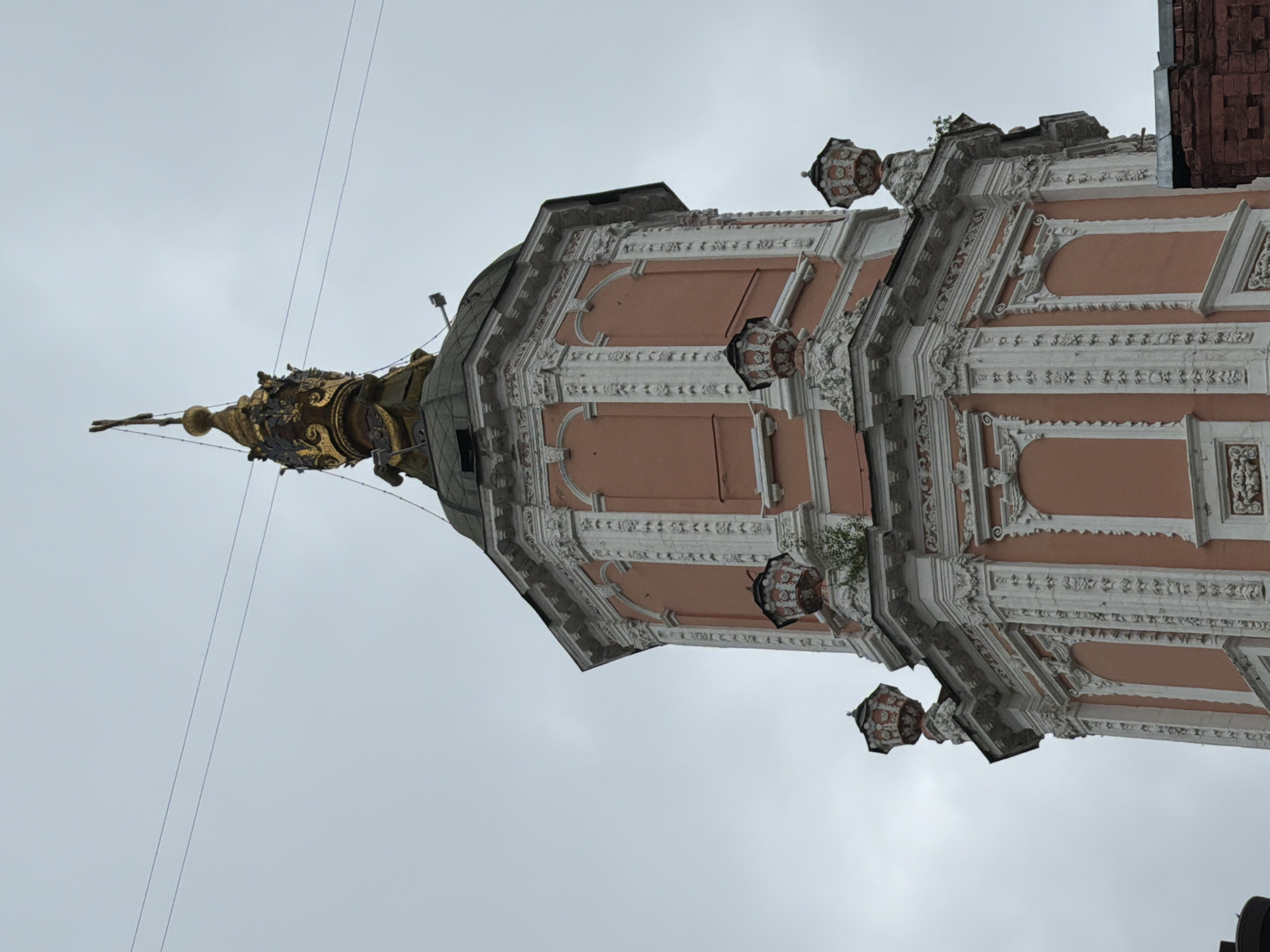 The image shows the Church of the Archangel Gabriel, also known as the Menshikov Tower, in Moscow, Russia. The church features an ornate Baroque style with pink facades trimmed with white architectural details. It has multiple tiers and a distinctive spire topped with a gilded figure. The sky is overcast.