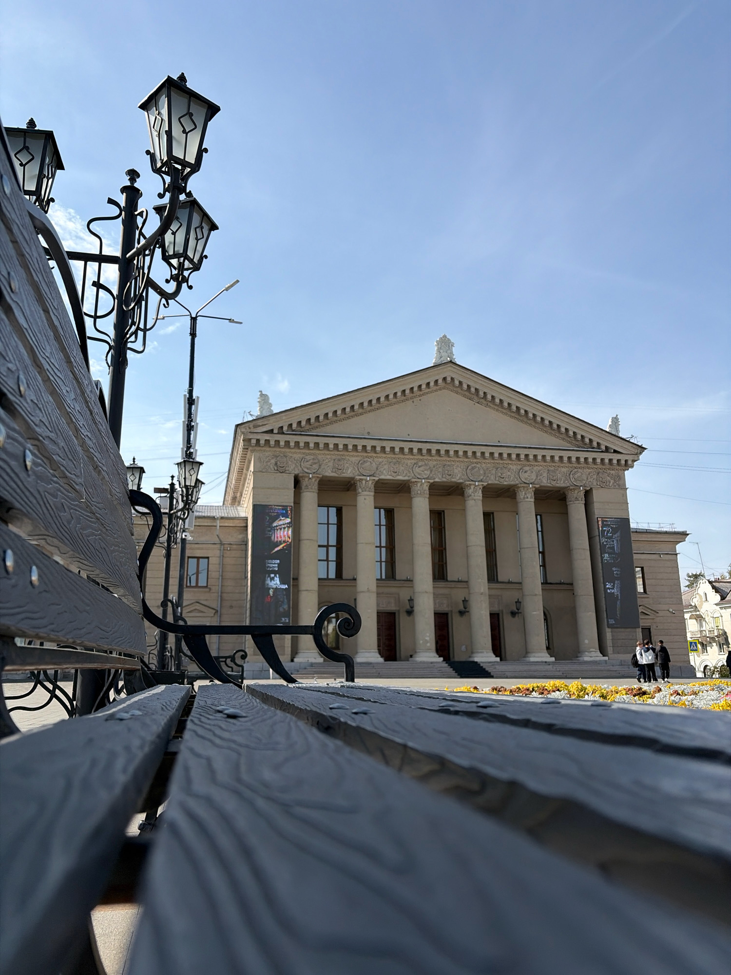 The image shows a low-angle shot of a street lamp and a park bench in the foreground, with the Angarsk Drama Theater in the background. The theater is a large, neoclassical building with a colonnade and a pediment. The sky is clear and blue. There are a few people walking in front of the theater. The bench is made of dark wood and has black metal legs and armrests. The street lamp is made of black metal and has three lights. The photo appears to have been taken on a sunny day.