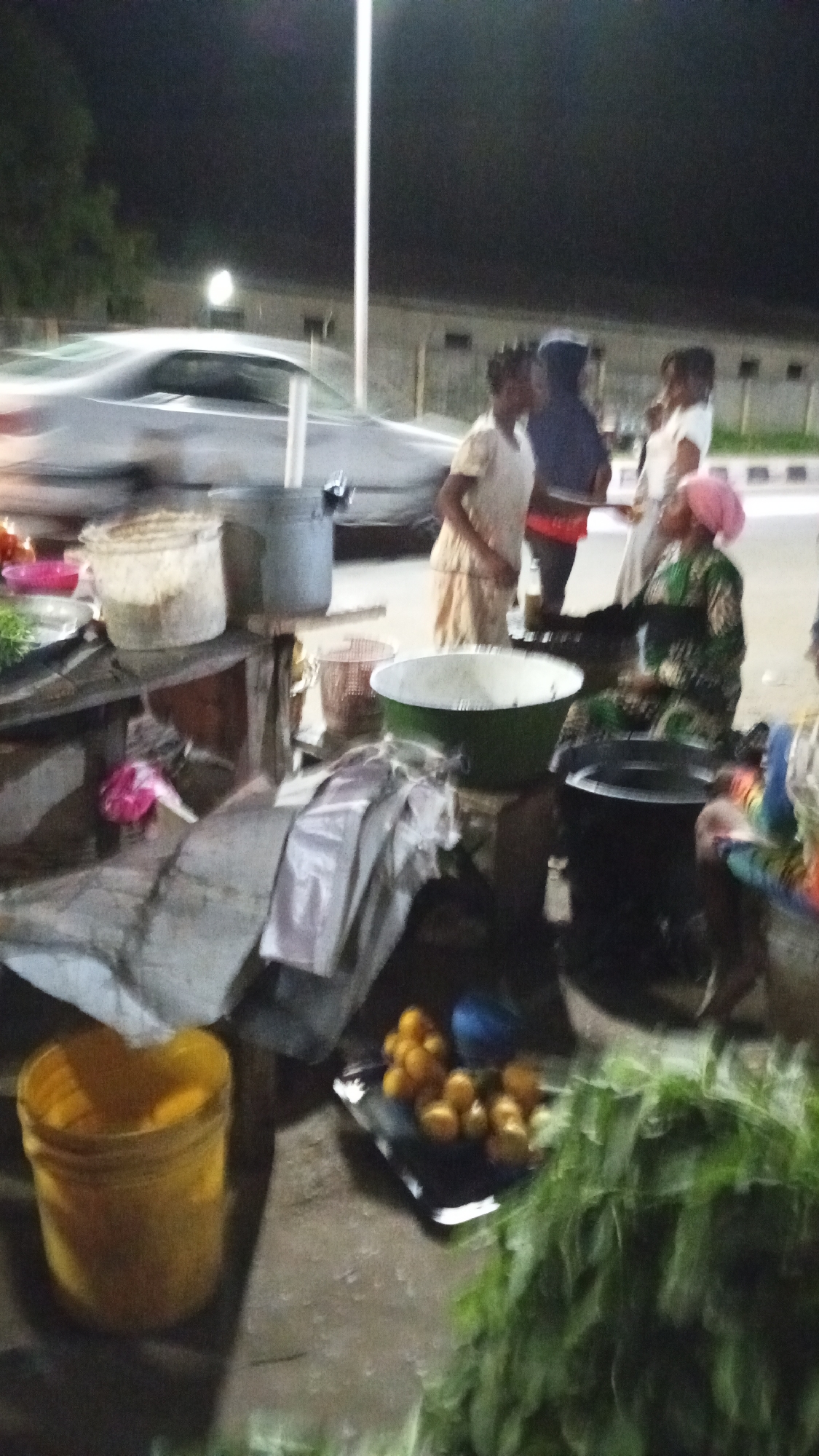 Captured at night, the image portrays a roadside market in Adankolo, Nigeria. A woman with a pink headscarf and patterned dress sits attending to customers. Other individuals stand nearby, engaged in conversation. Several containers and cooking equipment sit on a table, including a green basin, a black pot, and various buckets. A black tray holds oranges. A car drives by in the background. Street lights illuminate the scene. Green plants and miscellaneous items are strewn about.