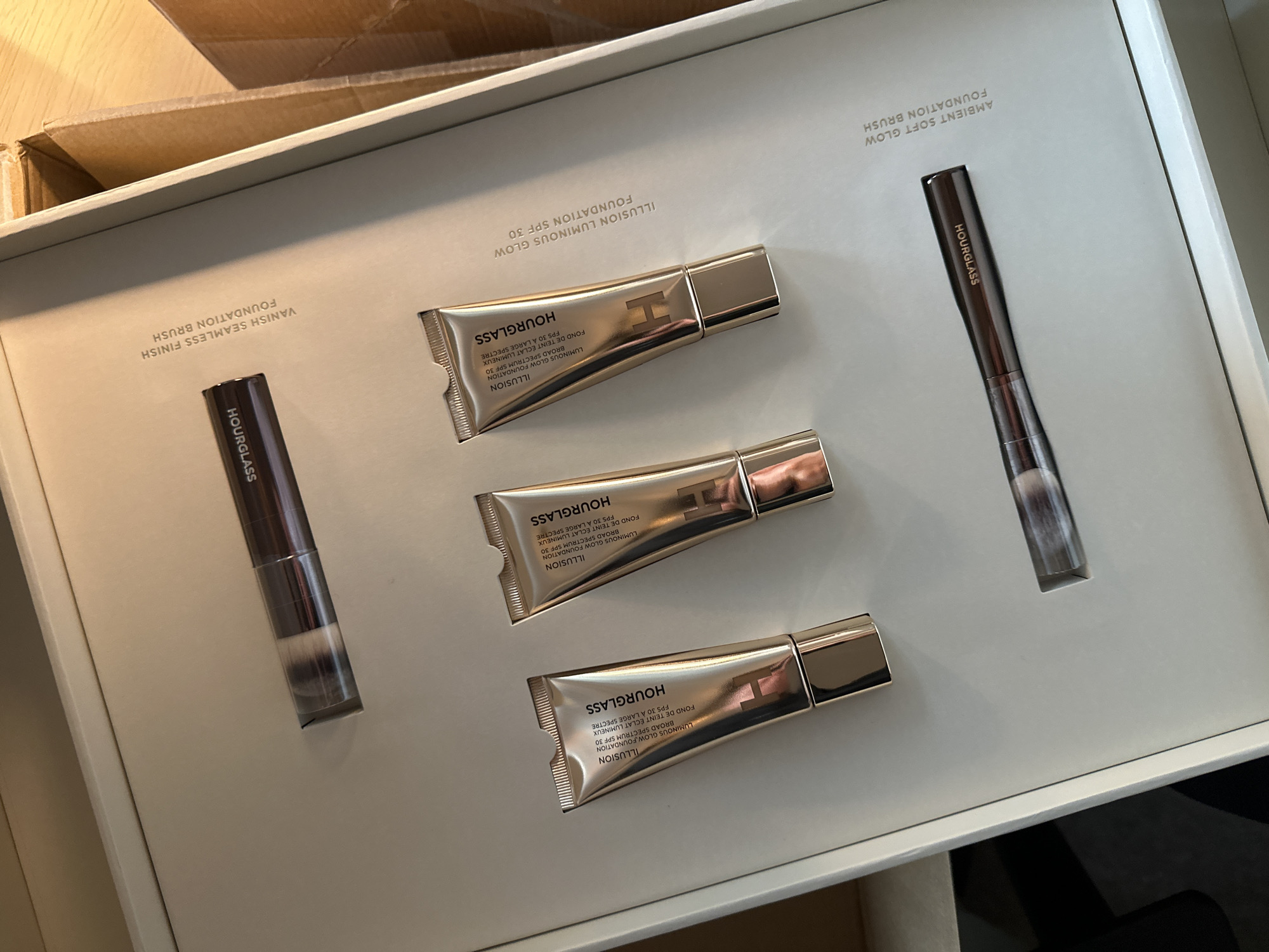 A close-up shot displays an open Hourglass cosmetics gift set, neatly arranged within a white box. The set contains three tubes of Hourglass Illusion Luminous Glow Foundation SPF 30, each with a metallic silver finish. Positioned on either side of the tubes are two Hourglass makeup brushes: the Ambient Soft Glow Foundation Brush and the Vanish Seamless Finish Foundation Brush. The text on the packaging identifies the products, with details about the foundation's SPF and luminosity. The overall scene is a high-angle, product-focused shot.