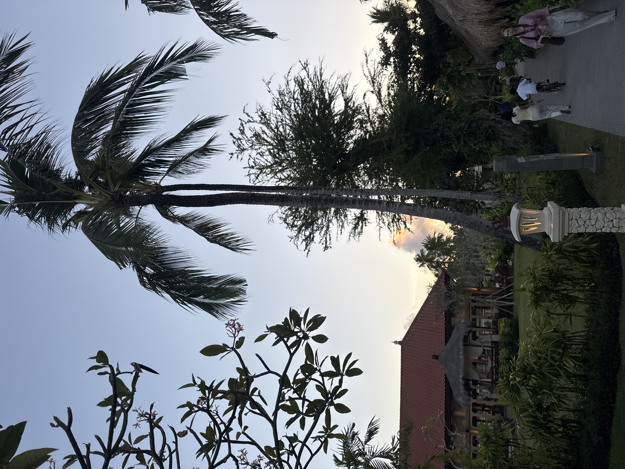 The image depicts a pathway lined with lush greenery and palm trees, leading towards a traditional building with a red tiled roof in Kuta Selatan, Indonesia. The sky is a soft blue, transitioning into a warm sunset glow in the distance. Several people are visible on the pathway, some riding bicycles and others walking. A woman in a pink shirt and white pants walks towards the foreground, carrying a bag. To the right, a thatched-roof structure is partially visible. The overall atmosphere suggests a serene evening in a tropical resort setting.