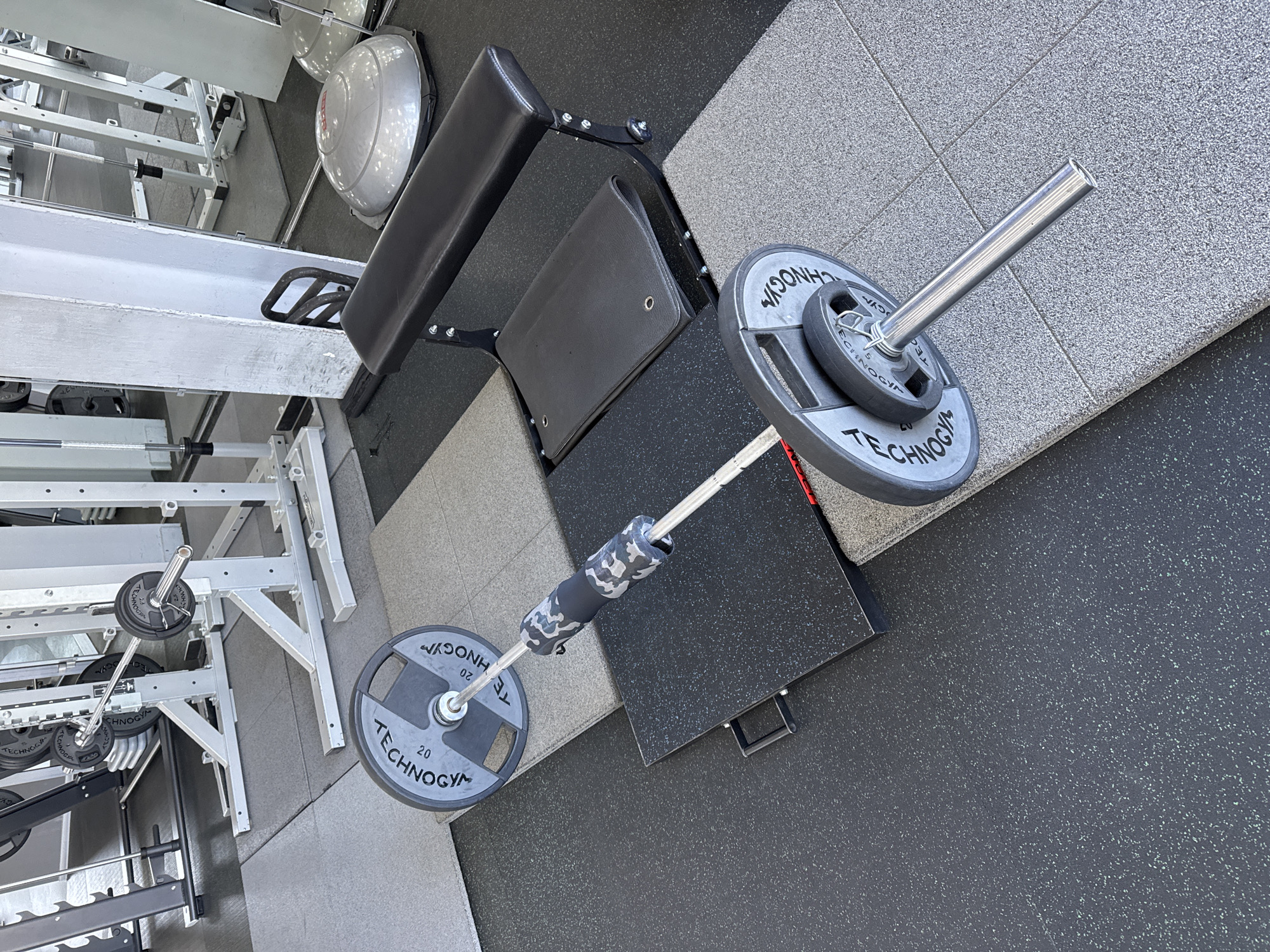 The image depicts an indoor gym setting, likely in Belorechensk, Russia, focusing on exercise equipment. The main subject is a barbell lying on a speckled rubber floor. It is equipped with multiple weight plates, prominently featuring "TECHNOGYM" branding. Visible weight denominations include "20" kg on larger plates and "2.5" kg on a smaller plate. A black and white camouflage-patterned squat pad is positioned on the center of the barbell.
To the right of the barbell is a black, adjustable weight bench with upholstered padding. In the background, a silver-colored medicine ball or balance trainer is partially visible. The gym floor is covered with two types of rubber tiles: a darker, more densely speckled black/grey, and a lighter, less speckled grey. Various other pieces of gym equipment, including metal weight racks and structural components of fitness machines, are visible in the background, particularly on the left side of the frame. No people are present in the image, and no activity is occurring; the equipment appears to be set up and ready for use. The lighting suggests a well-lit indoor environment.
