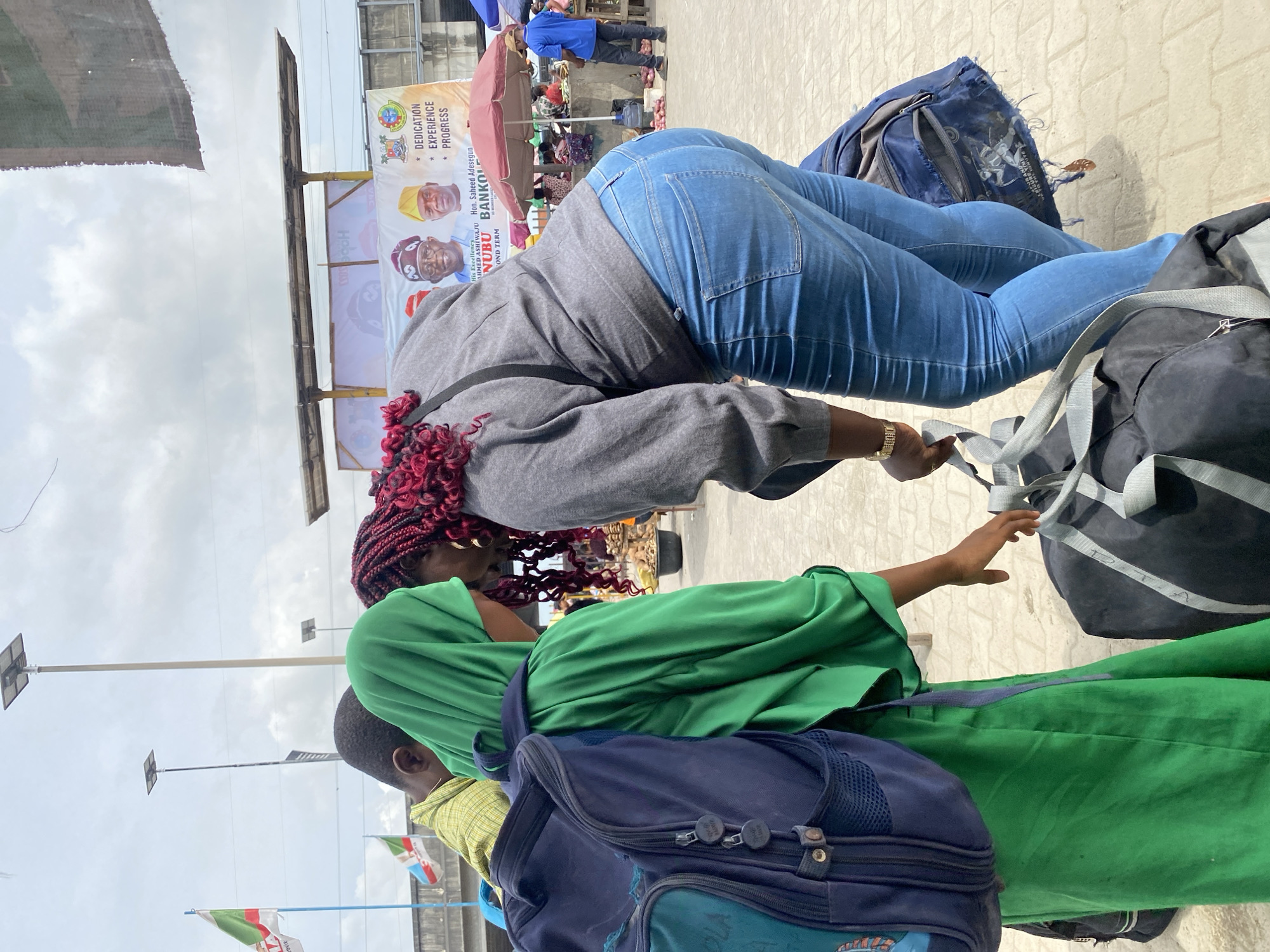 The image depicts an active street scene in Lekki, Nigeria, under an overcast sky during daytime. The foreground is dominated by several individuals and their luggage, suggesting movement or travel.
A woman with reddish-brown braided hair, wearing a grey sweatshirt and blue jeans, is seen from behind, bent over a large black duffel bag, seemingly adjusting or retrieving something. Another blue bag rests near her feet. Adjacent to her, a person in a bright green garment is partially visible from the back, accompanied by a blue backpack. A child or young person in a yellow plaid shirt is also seen, dressed in a green outfit.
In the mid-ground and background, the setting reveals a bustling urban environment, likely a market or commercial area, with people, market stalls, and a red umbrella. Overhead, streetlights equipped with solar panels are visible against the cloudy sky. Prominent political billboards stand in the background, featuring portraits of individuals. The text on these banners includes "DEDICATION EXPERIENCE PROGRESS," "His Excellency AHMED ASHIWAJU," "Hon. Saheed Adesegun," "NUBU BANKOLE," and "OND TERM," likely referencing a "Second Term." Flags displaying green, white, and red colors, consistent with Nigerian political party flags, are also present. The ground appears paved, characteristic of an urban walkway. The scene portrays a slice of everyday life in a vibrant Nigerian city.