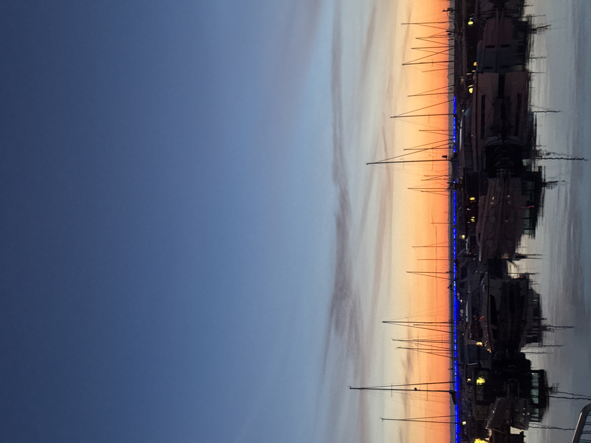 The image depicts a serene harbor scene at dusk, with the sky transitioning from a deep blue at the top to warm hues of orange and pink near the horizon, where several sailboats are docked. The water reflects the colorful sky and the silhouettes of the boats, creating a tranquil atmosphere. Some of the boats have lights on, casting a warm glow on the water. A thin, bright blue line of lights runs along the waterfront behind the boats. The boats are mostly modern yachts with tall masts. The scene is set in the city of Tallinn, Estonia. There are no people visible in the image.