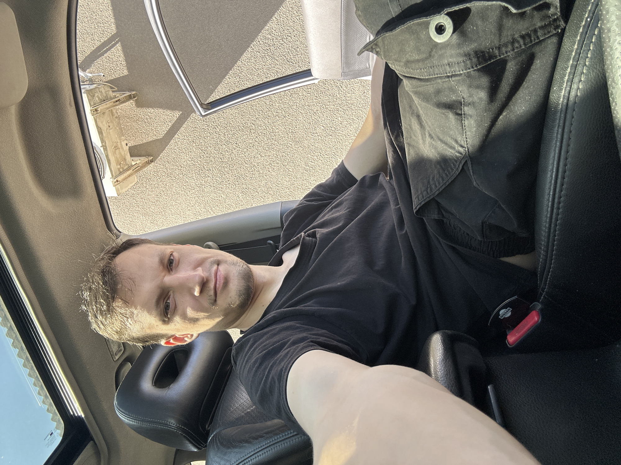 The image shows a man sitting in the driver's seat of a vehicle, possibly a Nissan given the logo on the seatbelt buckle. He is wearing a black t-shirt and dark cargo pants. The car has a sunroof open, and the exterior wall of a building, possibly with an air conditioning unit mounted on it, is visible through the open door and window. The lighting suggests it is daytime and sunny. There is no indication of Shatava, Ukraine, in the image itself.