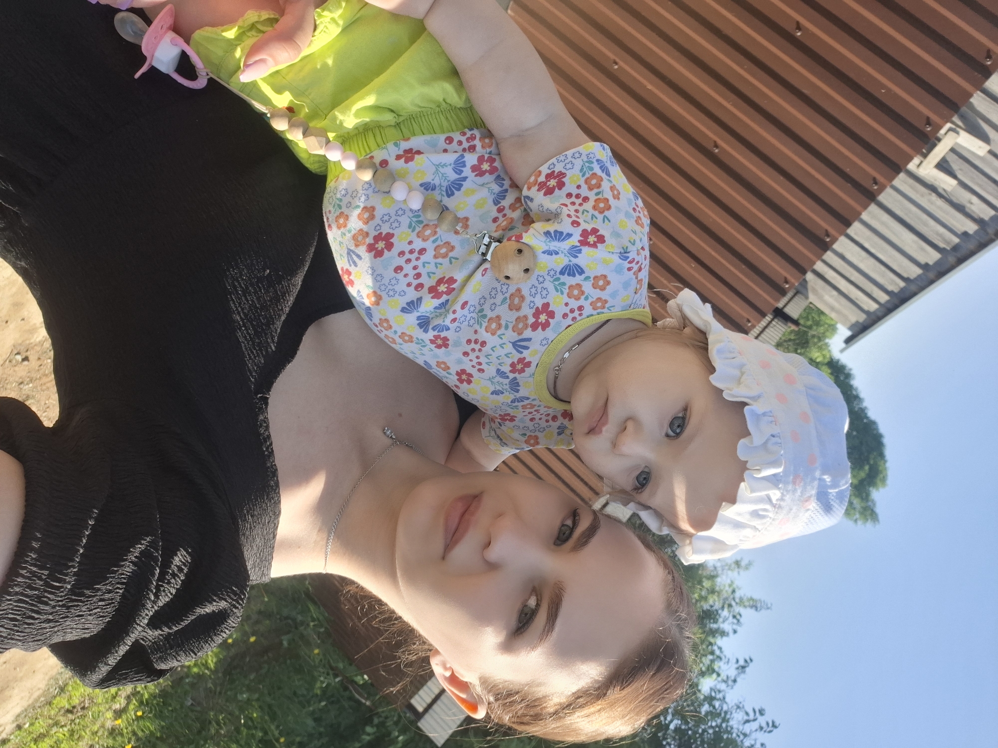 Here is a description of the image:
A woman and a baby are posing for a selfie outdoors. The woman has fair skin, light brown hair pulled back, and is wearing a silver necklace and a black top. The baby, dressed in a floral shirt, yellow shorts, and a white bonnet with pink polka dots and a ruffled brim, is nestled against her chest. A wooden beaded pacifier clip is attached to the baby's outfit. The background shows a metal structure, possibly a roof or a wall, along with glimpses of greenery and a blue sky. They are likely in the city of Nizhnyaya Gar', Russia.