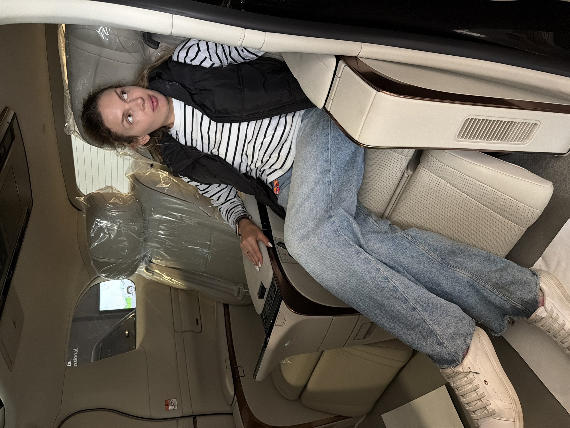 A woman is lying inside the back of a car. She is wearing a black and white striped long-sleeved shirt, a black vest, and blue jeans. Her feet are shod in white sneakers. The car's interior is beige with wood trim. The car's seats are covered in plastic. The setting is likely Podgorica, Montenegro, based on possible geolocation information associated with the original image file. The image suggests a staged or promotional setting rather than a natural, unposed scenario.