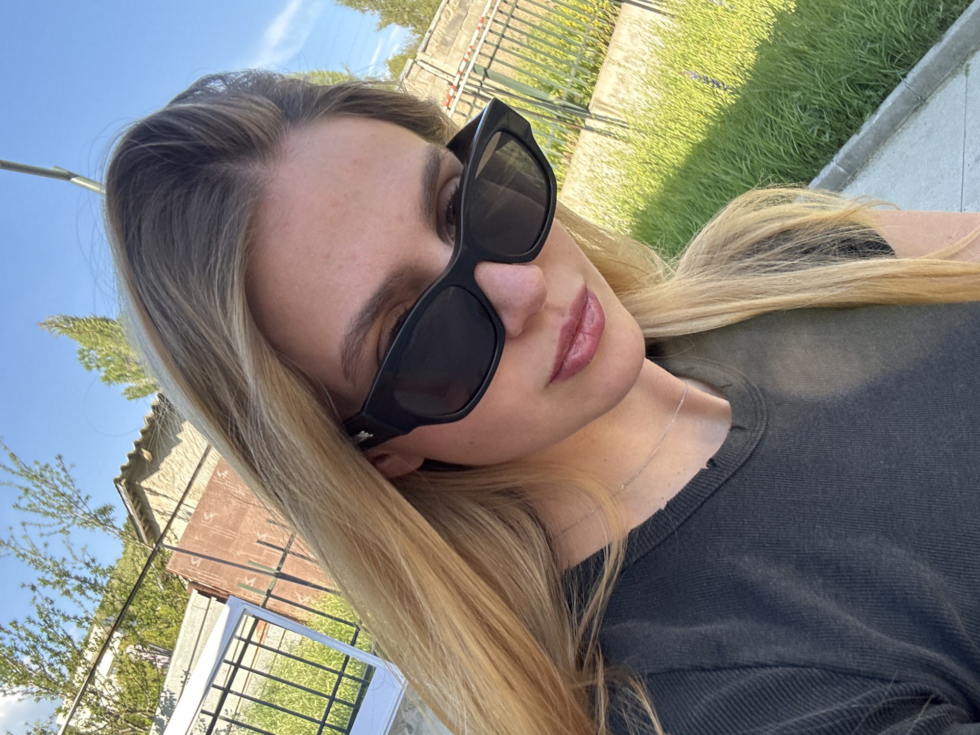 A medium-shot portrait features a young woman with blonde hair, wearing black sunglasses, and a dark gray crewneck shirt. She is positioned outdoors on what appears to be a sunny day, with a blue sky partially visible.
Behind the woman, the background includes a mix of urban and natural elements. There is a building with a weathered roof and fencing, along with lush green grass. A concrete pathway or sidewalk is also present.
Given the context of the city location, it is plausible that the photo was taken in an urban park or a residential area on the outskirts of Tbilisi, Georgia. The background suggests a blend of built structures and greenery common in many parts of the city.