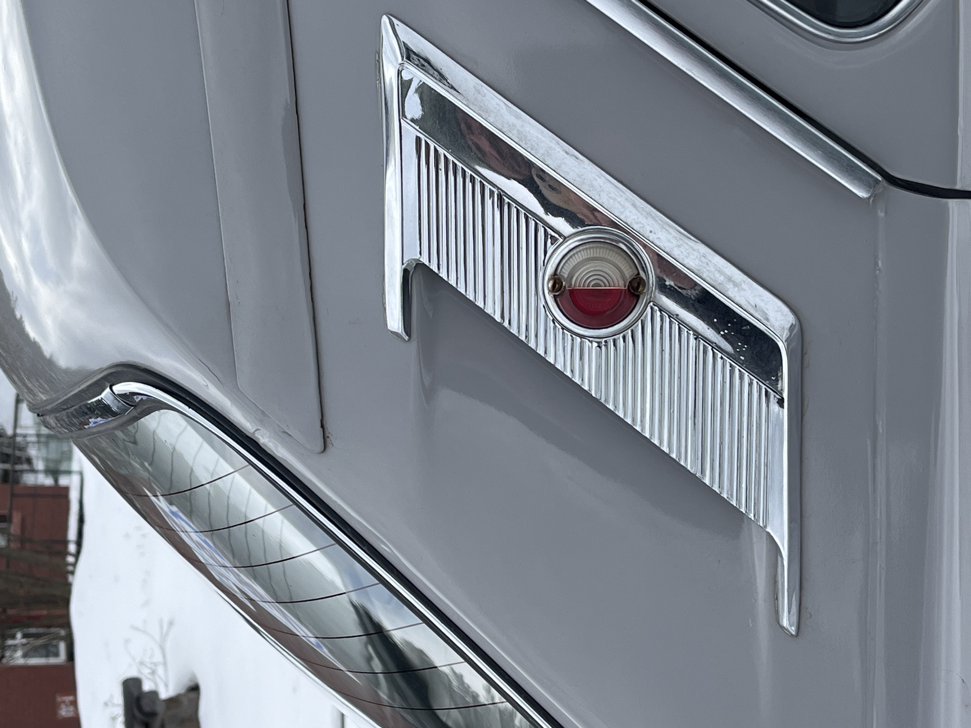 A close-up shot depicts the taillight and chrome trim of a gray classic car. The taillight, encircled in chrome, features a red lens. The chrome trim has a series of vertical ridges and is shaped like an arrow. The car is likely outdoors given the blurry scene reflected in the chrome and the presence of snow and partial buildings in the background. The location in the city of Unknown, Unknown cannot be determined.