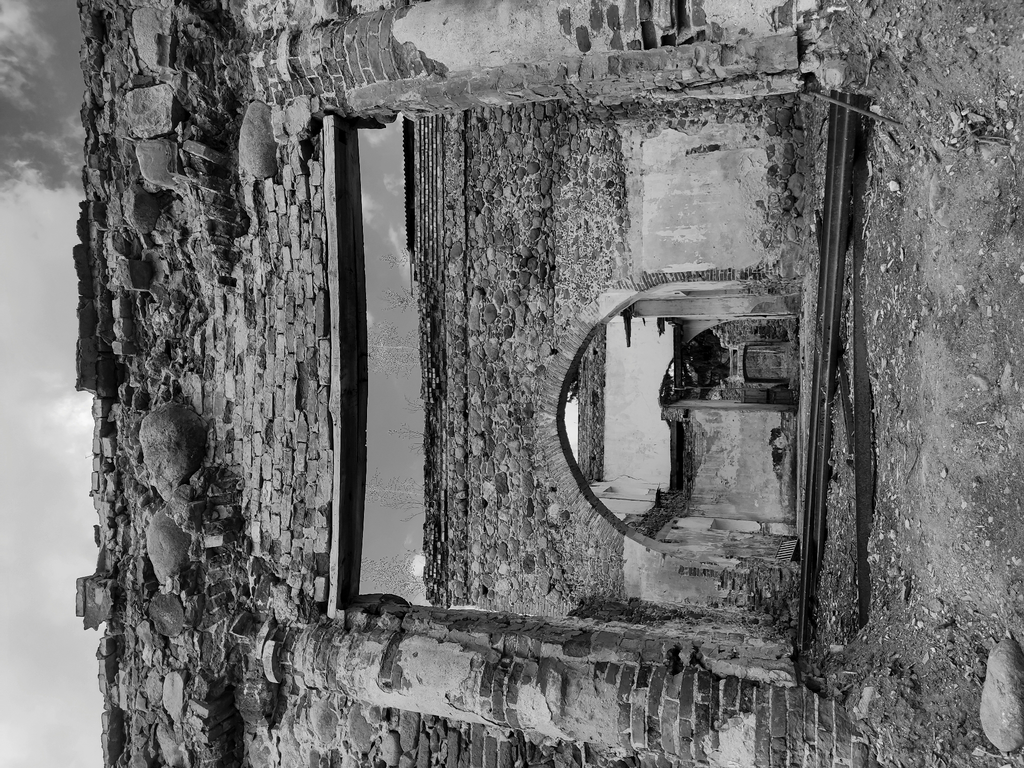 The image presents a black and white view of a ruined structure in Жалудоцкі сельскі Савет, Belarus. The building appears to be constructed from a combination of brick and stone. There are visible openings, including a window and an archway, which reveal further interior deterioration. The environment is marked by rubble and debris.