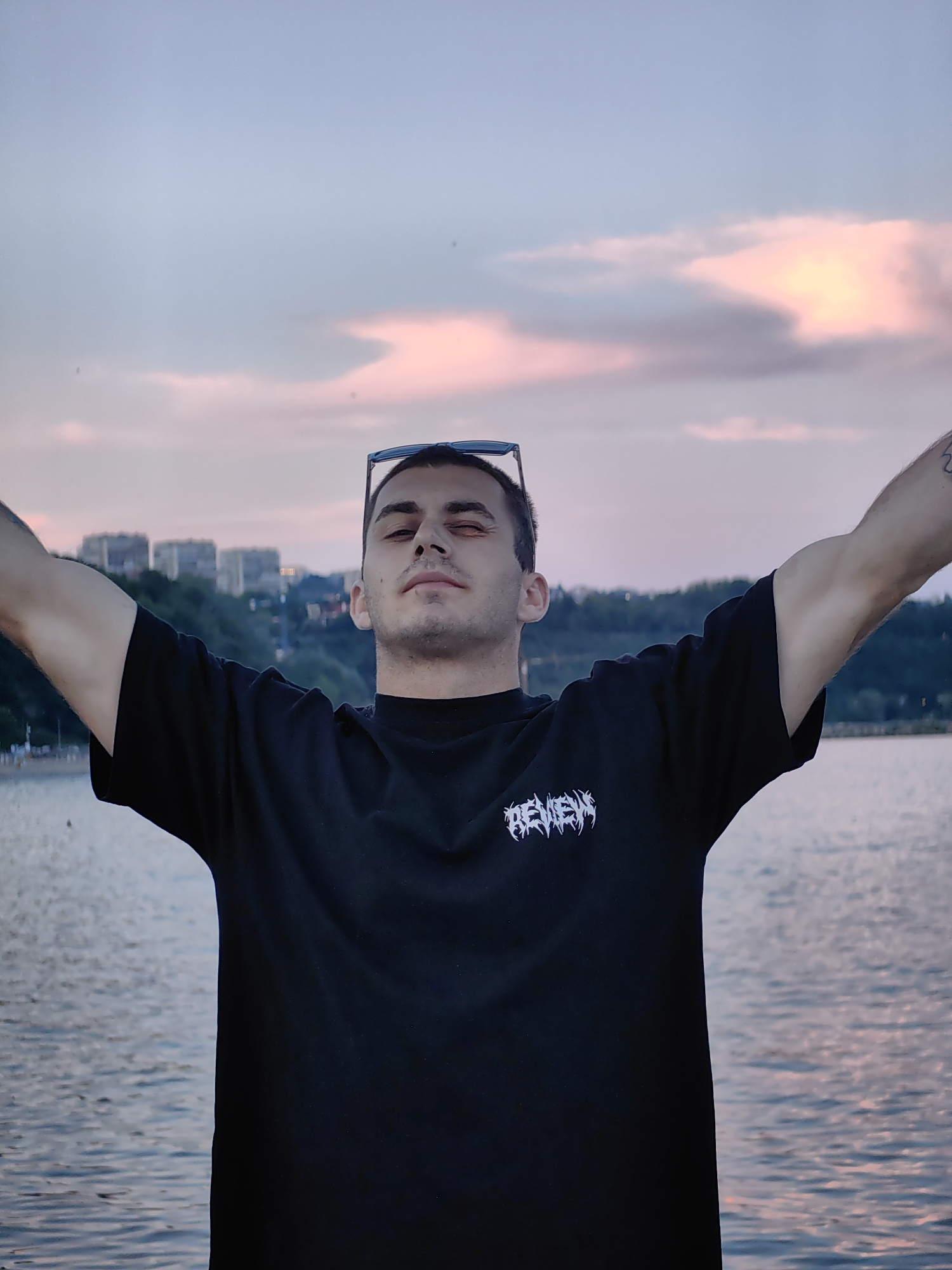 A young man with dark hair stands with his arms raised and eyes closed against a twilight sky. He wears a black t-shirt with a white graphic logo on the left chest and sunglasses perched on his head. The background features a body of water with ripples, and across the water, a city skyline is visible with buildings and trees lining a hill. The sky is a gradient of pale blue and soft pink, indicating the setting or rising sun.