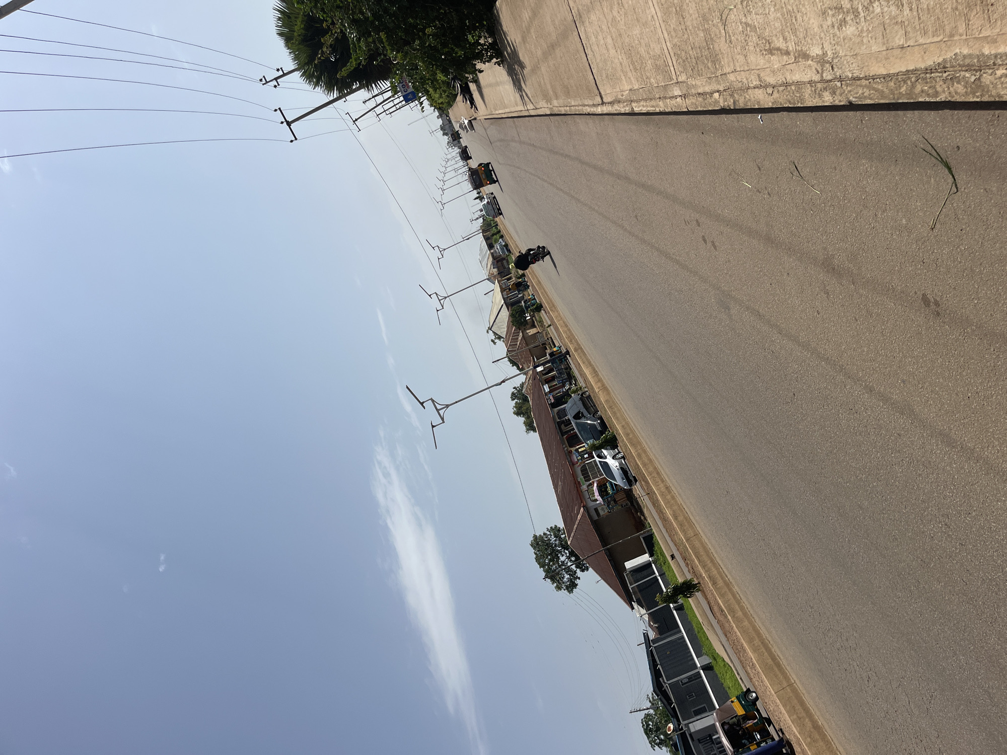 This is a daytime, eye-level, outdoor, eye-level shot of a street in Bukuru, Nigeria. The image is angled from the right side of the road, looking down the length of the asphalt road. Several vehicles are visible on the road, including a tuk-tuk, a motorcycle, cars, and possibly a van. Buildings line the left side of the road, appearing to be shops or residences with corrugated metal roofs and some covered stalls. Modern streetlights with solar panels are installed along the road. Utility poles and wires are visible on the right side of the frame, along with dense green foliage from trees. The sky is mostly clear with some scattered clouds. The road has painted lane markings and a concrete sidewalk on the right. The scene depicts a typical urban street environment with ongoing traffic and commerce.