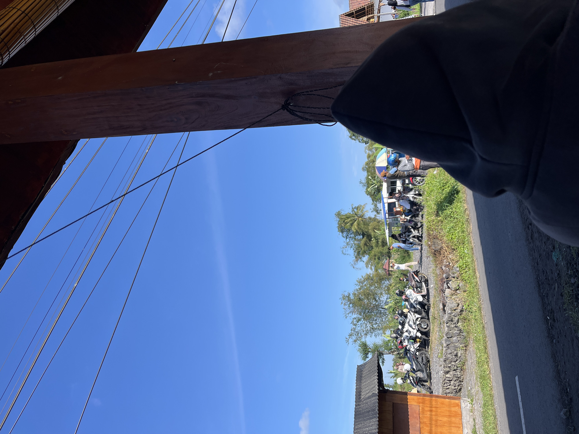The image depicts an outdoor scene in Oro-oro Ombo, Indonesia, under a clear blue sky with scattered clouds. In the foreground, the back of a person wearing a dark hooded sweatshirt is visible, obscuring their face. To the left, a small wooden building with a dark, textured roof stands adjacent to a stone wall. A line of several parked motorcycles, mostly white and black, are visible in front of the stone wall. Several individuals are standing near the motorcycles, some appearing to be in conversation. Further in the background, there are trees and vegetation, including bamboo, and a glimpse of buildings with tiled roofs. Power lines are stretched across the upper portion of the frame, supported by a large wooden pillar on the right. A road runs across the bottom of the image.