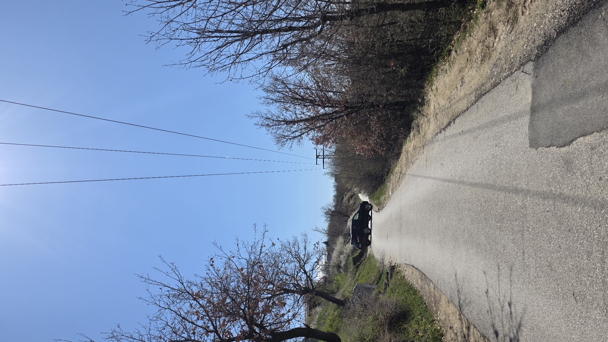 The image shows a narrow paved road curving through a rural area near Elenovo, Bulgaria. A dark-colored hatchback vehicle is parked on the road, facing away from the viewer. The road is bordered by dry vegetation and trees with bare branches. Overhead power lines stretch across the sky. The sky is clear and blue. There are no people visible in the image.