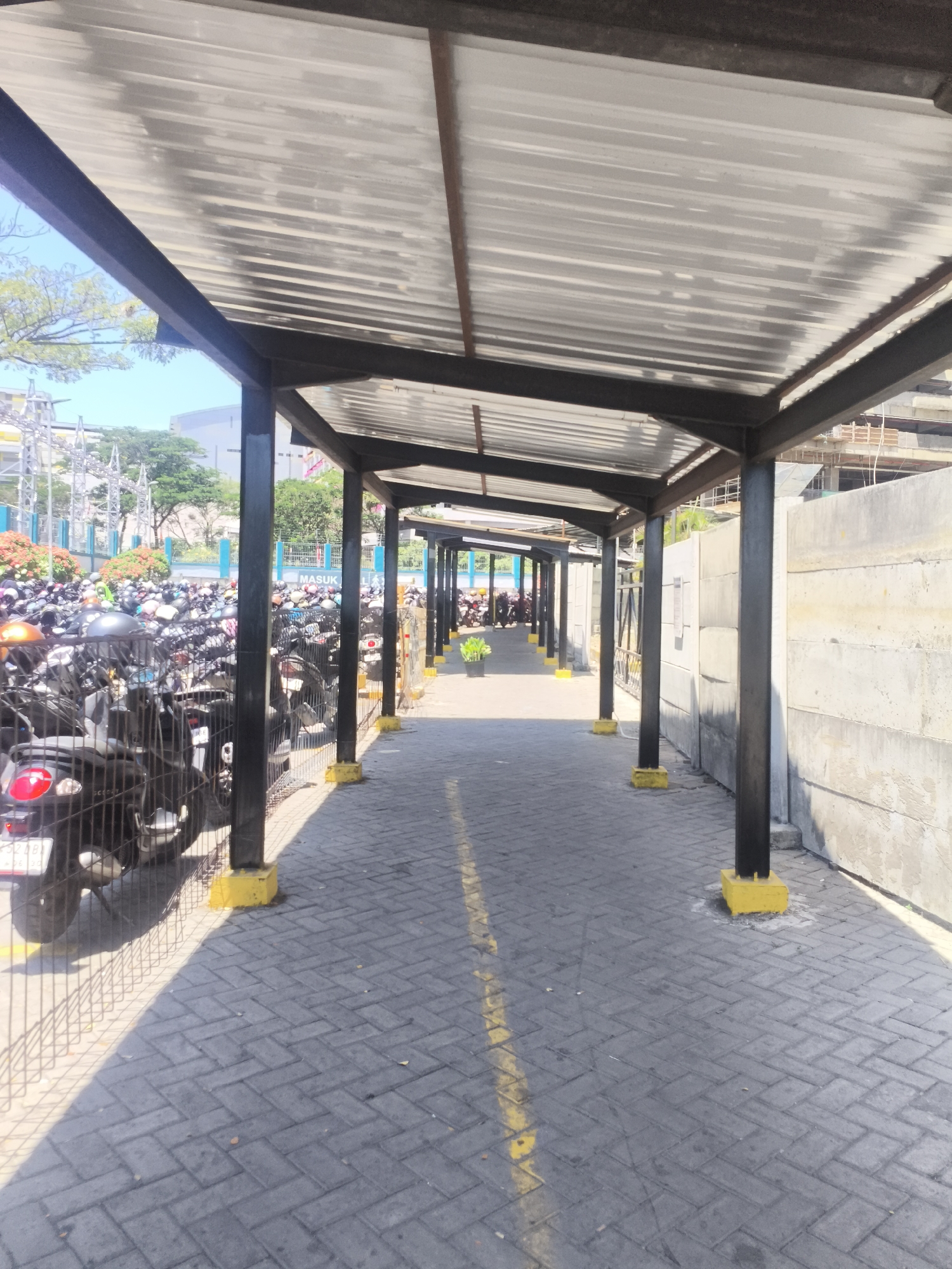 The image depicts an outdoor walkway with a corrugated metal roof supported by dark metal posts. The walkway is paved with gray interlocking bricks, and a yellow line runs down its center. On the left side of the walkway, a dense parking lot filled with numerous motorcycles is visible, separated by a wire fence. The motorcycles are of various colors and models. Some trees and foliage can be seen beyond the parking lot, along with buildings in the background, suggesting an urban environment. A person wearing a yellow vest is walking in the distance along the covered walkway. The right side of the walkway is bordered by a rough-textured concrete wall. Sunlight casts strong shadows across the paved surface, indicating a bright, sunny day. The scene is consistent with a covered walkway in a parking area or public space in Surabaya, Indonesia, where such covered walkways are common for protection from the sun and rain.