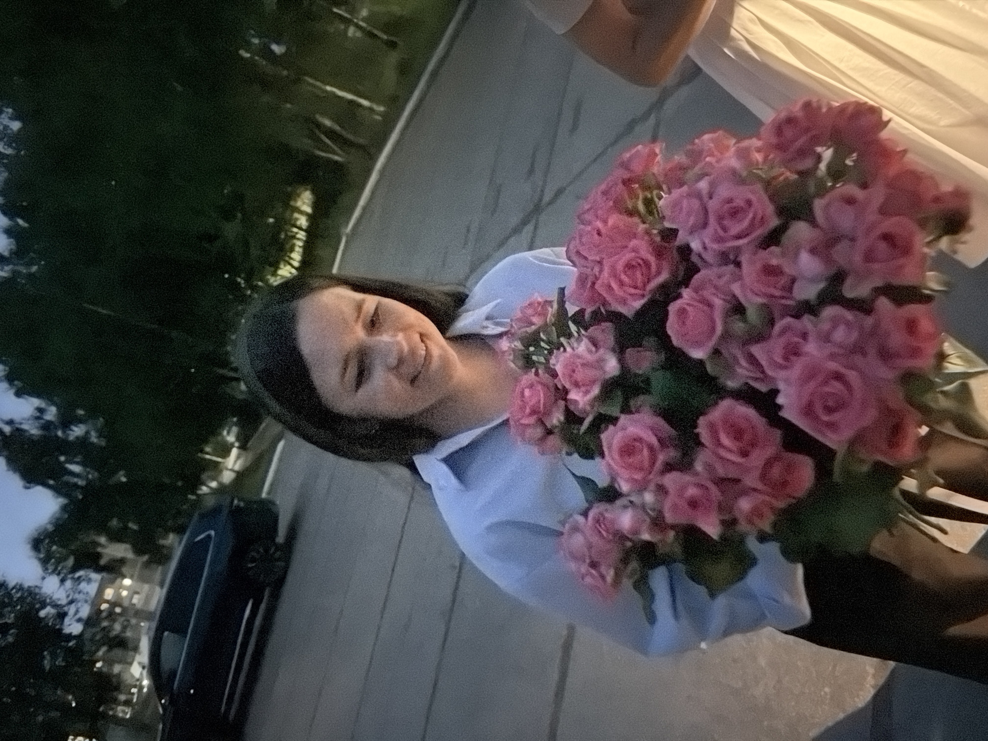 A young woman with dark hair smiles faintly as she holds a large bouquet of pink roses. She is wearing a light blue collared shirt and a black garment. The bouquet is held in front of her, partially obscuring her body. To the right of the woman, a person in a light-colored dress is partially visible. The background shows trees and a dark-colored SUV parked on an asphalt surface, possibly a street or parking lot. The lighting suggests it is dusk. The scene appears to be outdoors in a residential or urban area.