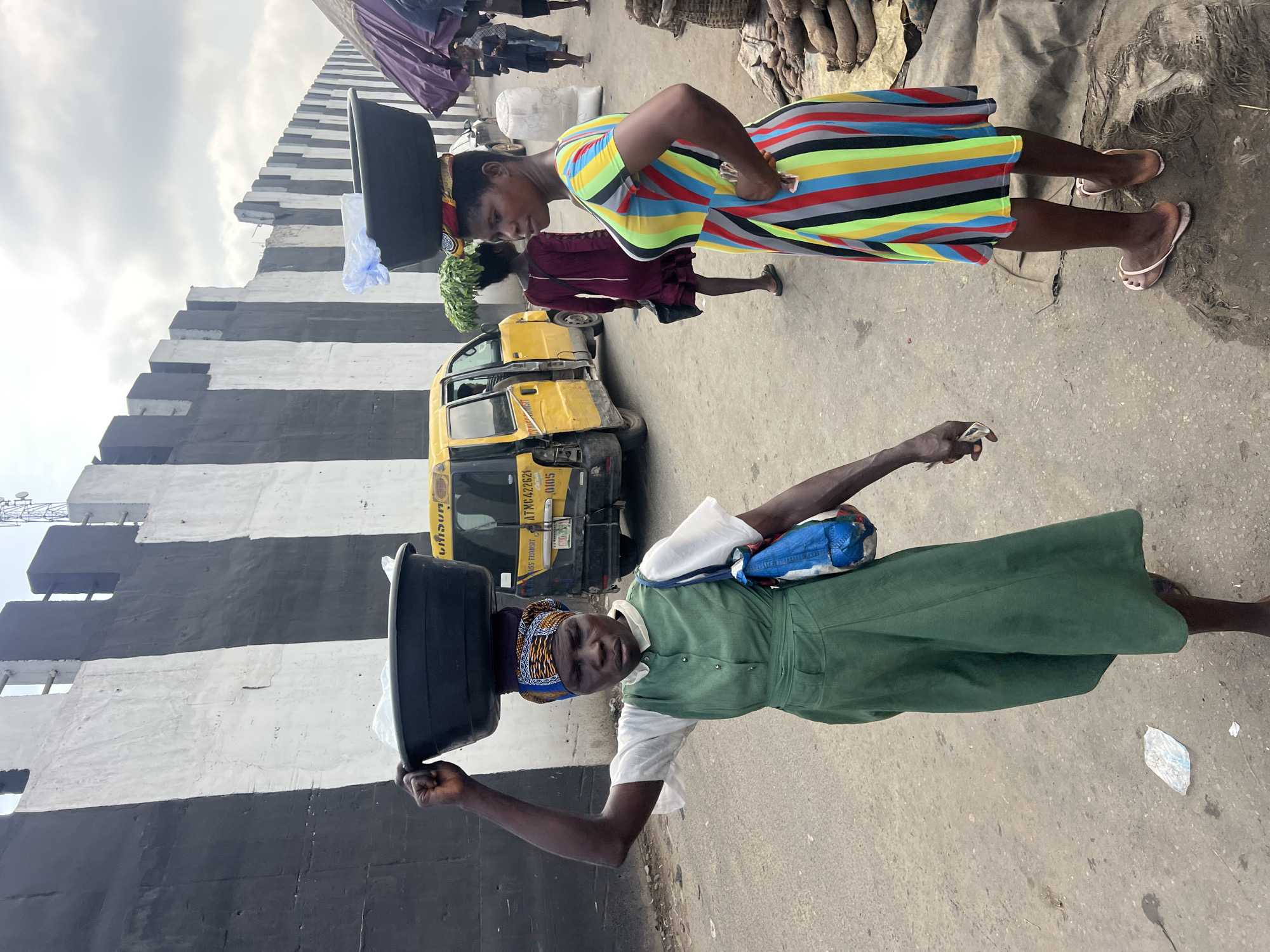 In Nkpor, Nigeria, two women are walking down a street, each carrying a large basin on their head. The woman in the foreground, on the left, wears a green dress over a white blouse and a colorful headscarf. She carries a black basin containing white plastic bags. To her right, another woman wears a brightly striped dress. She carries a black basin with produce, possibly leafy greens, peeking out from under it, balanced on a colorful cloth. In the background, a yellow commercial van is parked, and other people are visible walking or standing. The street is paved with asphalt, and a distinctive black and white striped wall runs along the left side of the scene. The sky is overcast.