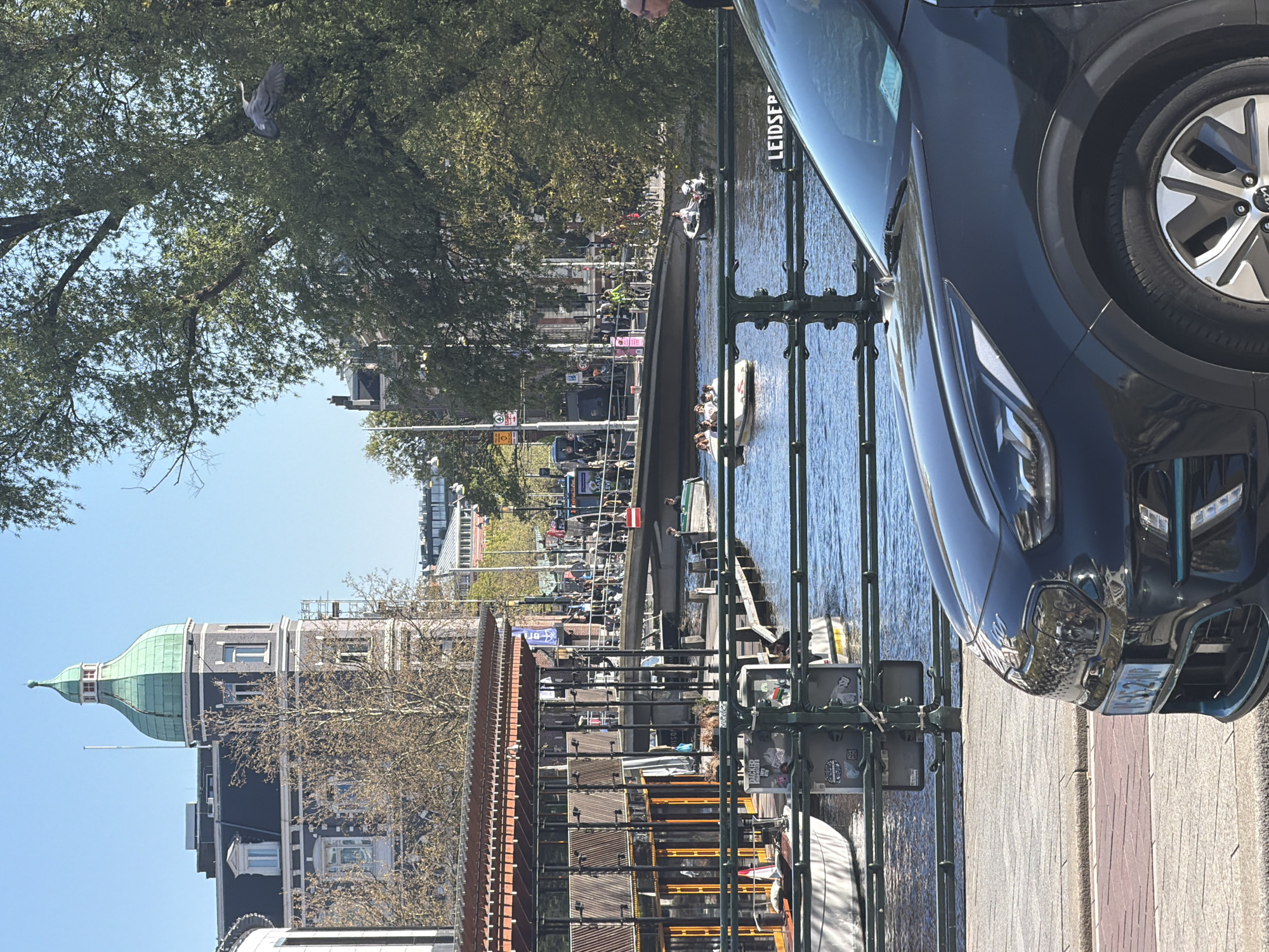 This media file captures a sunny daytime scene along a canal in Amsterdam, Netherlands. The perspective is slightly elevated, looking across the canal to the opposite bank and buildings.
In the immediate foreground, the side of a dark-colored SUV, possibly a Kia, is visible, parked on a paved area next to a dark green metal railing. The vehicle's front bumper, headlights, and a wheel are prominent, with a partial license plate displaying "K4S 2HP".
The midground features a clear blue canal with several small boats, some carrying people, suggesting leisure or tour activities. Along the canal's far bank, pedestrians are visible, and a sign affixed to the canal wall clearly shows the text "LEIDSEP," indicating proximity to a location such as Leidseplein or Leidsegracht.
The background is characterized by classic Amsterdam architecture, including a notable building topped with a distinctive green-domed cupola, set against a bright, clear blue sky. Lush green trees with full foliage line the canal banks, suggesting either spring or summer. Overhead, a large bird, possibly a heron, is captured mid-flight amongst the tree branches. The overall impression is one of a vibrant and pleasant day in the city.
