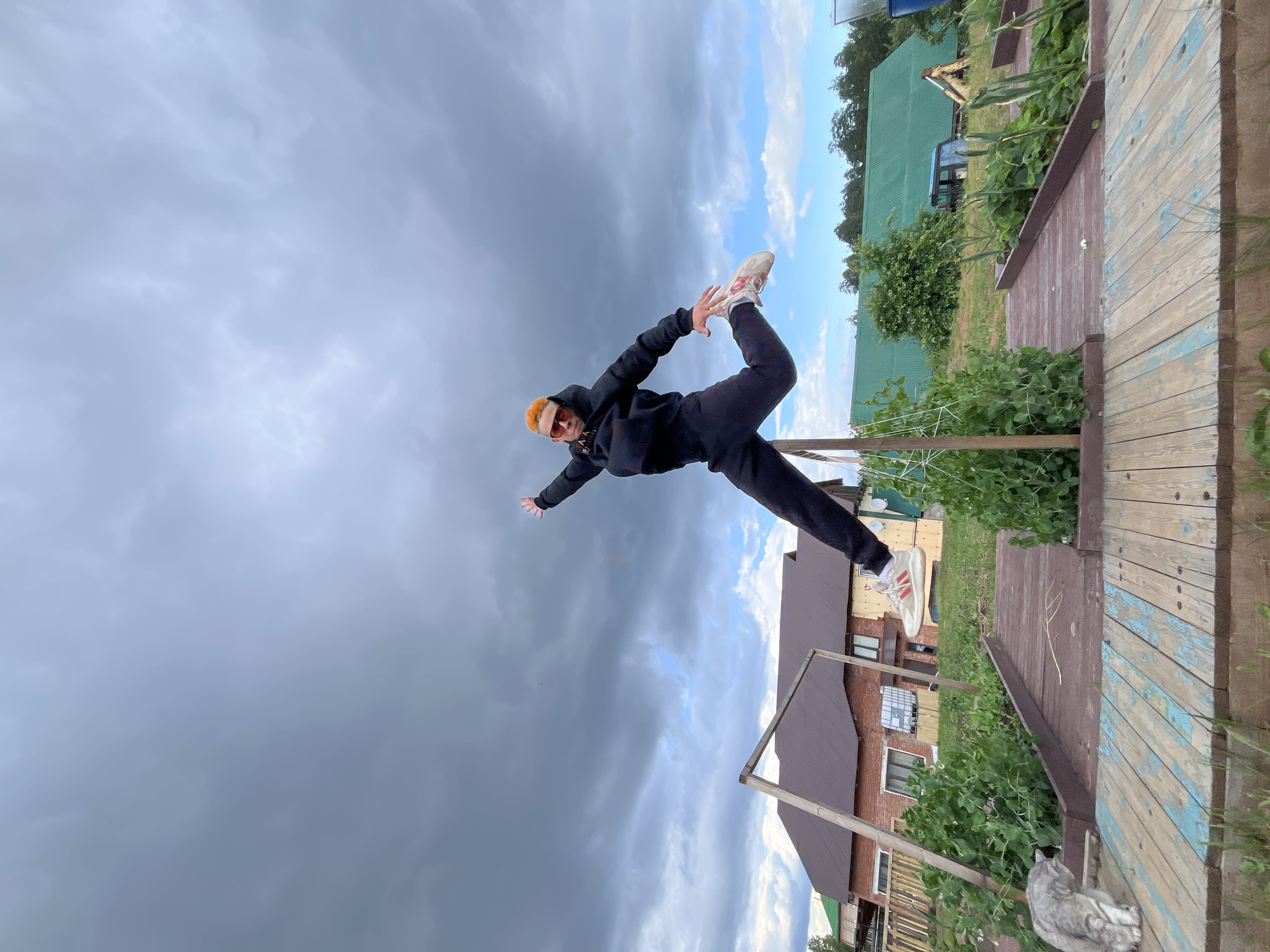 A person with bright orange hair, wearing sunglasses, a black hoodie, and black pants, is captured mid-air performing a yoga pose outdoors. They are balanced on one leg, with the other leg extended behind them, and their arms outstretched. The individual is wearing white athletic shoes with red accents.
The scene appears to be a rural or suburban setting. In the background, there are houses with brown roofs, green vegetation, and trees. A wooden structure, possibly a deck or walkway, is visible in the foreground and to the right. A light blue sky with scattered white clouds is visible, contrasting with darker, ominous storm clouds dominating the left side of the frame. This suggests a dynamic weather change.
The overall impression is one of athleticism and a moment of graceful movement against a dramatic sky. The location context places this scene in Chaikovsky, Russia. There is no visible text in the image.
