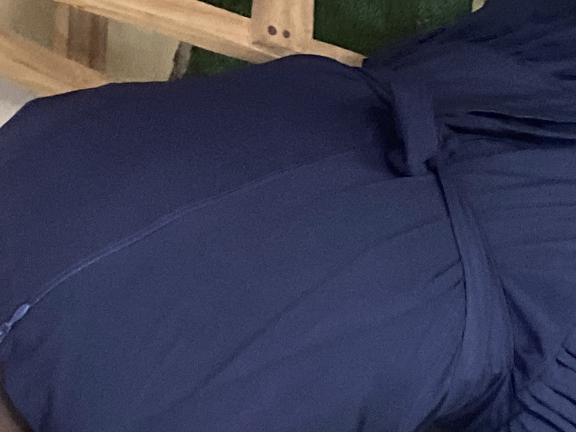 A close-up image shows a person wearing a dark blue dress with a sash tied around the waist. The dress appears to be made of a flowing material, possibly silk or satin, as it reflects light and shows soft folds and pleats. A zipper is visible on the upper left side of the dress. The background consists of a wooden structure, possibly part of a chair or furniture, and a patch of green, which could be grass or carpet. The overall setting suggests an indoor or outdoor environment where someone is dressed formally. The image does not provide any specific clues about the location within Jauro Yinu, Nigeria.