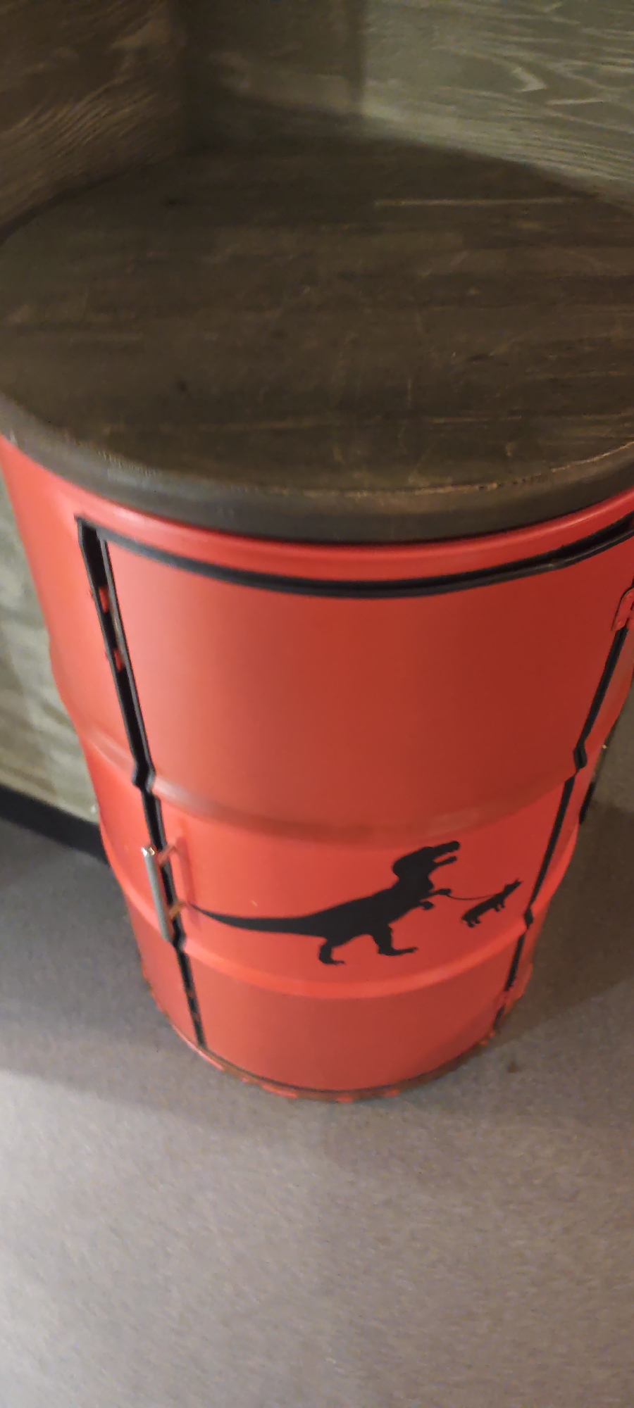 A red repurposed metal barrel with a wooden top sits on a gray carpet. A black silhouette of a Tyrannosaurus Rex walking a dog on a leash is painted on the side of the barrel. The barrel is segmented horizontally by metal bands. The location is Estosadok, Russia.