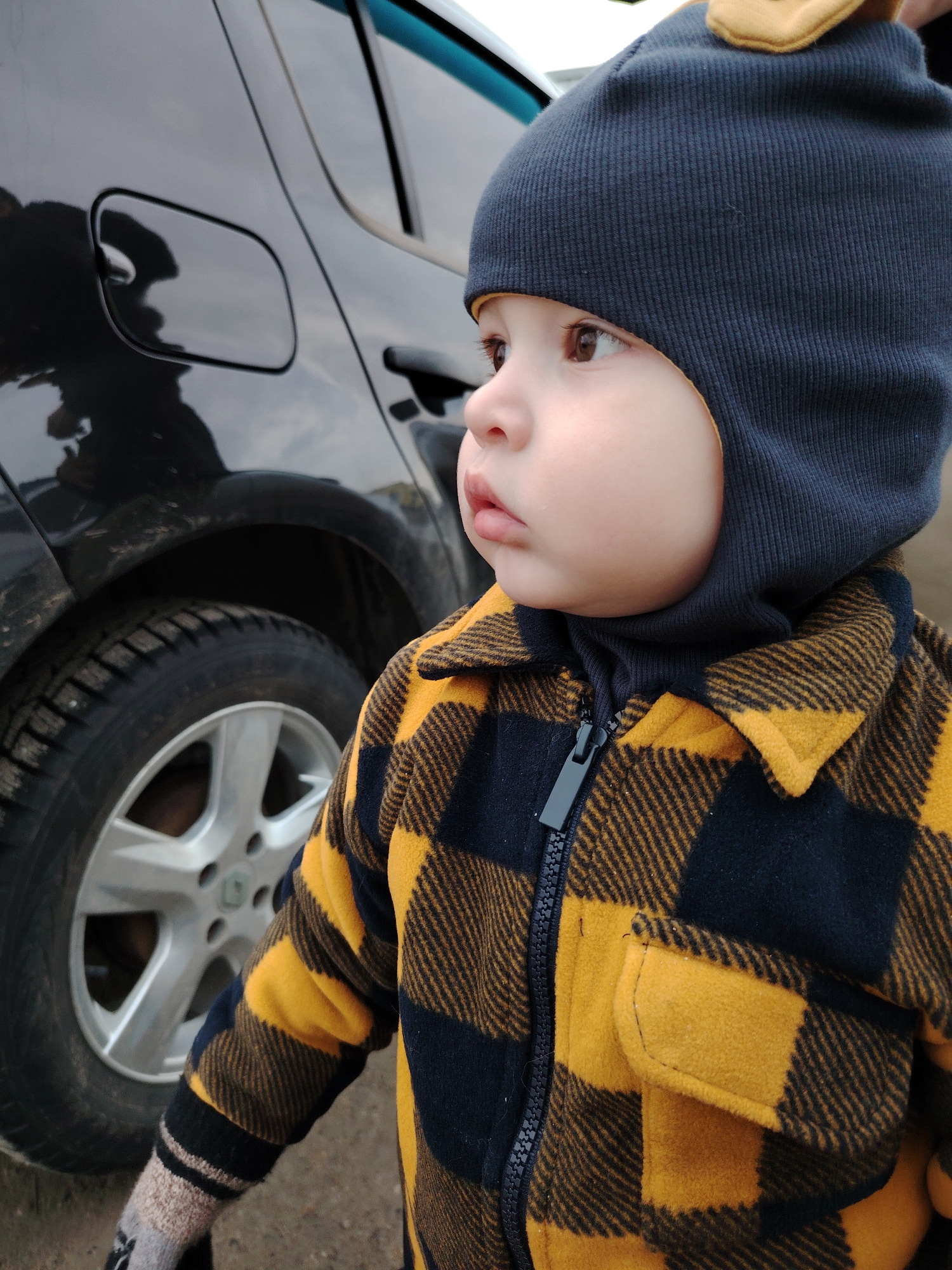 A young child stands outdoors, facing slightly to the right. The child wears a dark blue knit hat with small yellow accents, and a yellow-and-black plaid jacket. The jacket is zipped up. A black car is visible in the background, specifically the rear wheel and fuel door. The location is Chaikovsky, Russia.