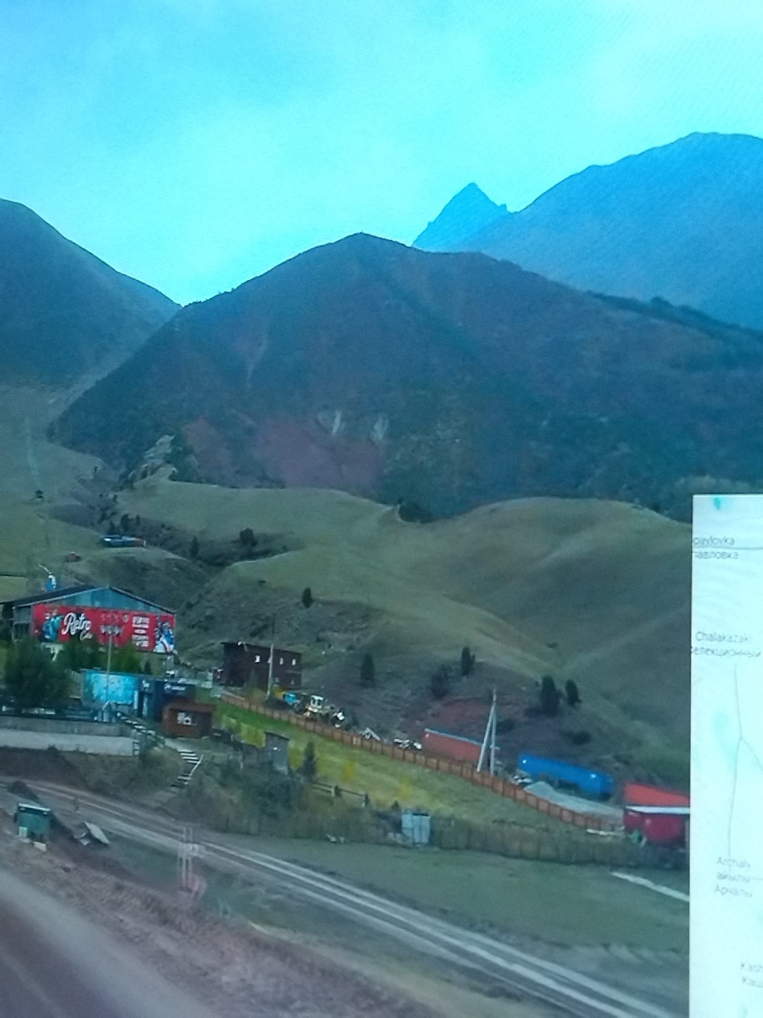 A wide shot captures a mountainous landscape with a clear blue sky. In the foreground, a cluster of buildings and structures are visible, some with red and blue roofs. A large red building with "Retro Cola" branding is prominent. Train tracks run through the scene, with a green container on the left side of the tracks. In the background, rolling green hills lead up to dark, shadowed mountains. A digital map is partially visible on the right side of the image, displaying text in a non-English language and a map of the area.