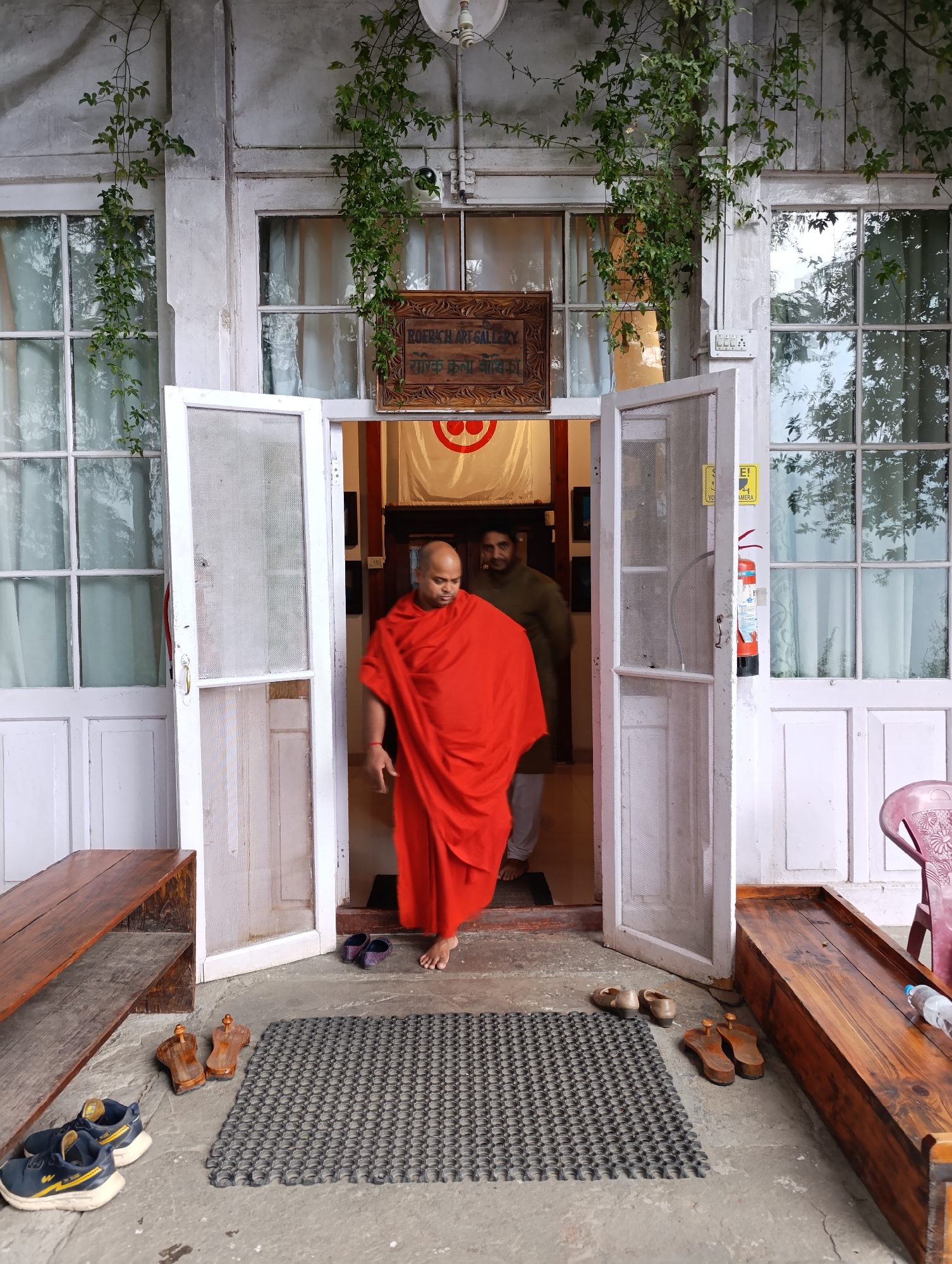 The image depicts an outdoor view of the entrance to a building in Naggar, India, likely an art gallery given the signage. The central subject is a person, possibly a Buddhist monk, dressed in flowing red robes, walking barefoot out of a white wooden screen door. Another person in a dark olive-green outfit is partially visible behind them inside the doorway.
The building features a weathered white facade with multiple paneled glass windows and white frames, adorned with lush green climbing vines. Directly above the entrance, a wooden sign clearly reads "ROERICH ART GALLERY" in English, with additional text in Devanagari script below it. A yellow sign affixed to the doorframe is partially visible, displaying "SE!" and "YOUR CAMERA," suggesting instructions or warnings about photography.
On the concrete ground in front of the entrance, several pairs of shoes and sandals are neatly placed, indicating a custom of removing footwear before entering. These include dark blue sneakers, two pairs of traditional wooden sandals (padukas), and dark-colored slippers. A black rubber doormat lies directly in front of the doorway. Wooden benches or low tables flank the entrance on both sides, with a plastic water bottle resting on the right-side bench. A red fire extinguisher is mounted on the wall to the right of the door, and a security camera and light switch are also visible on the building's exterior. The scene suggests a calm, culturally significant location during daytime.