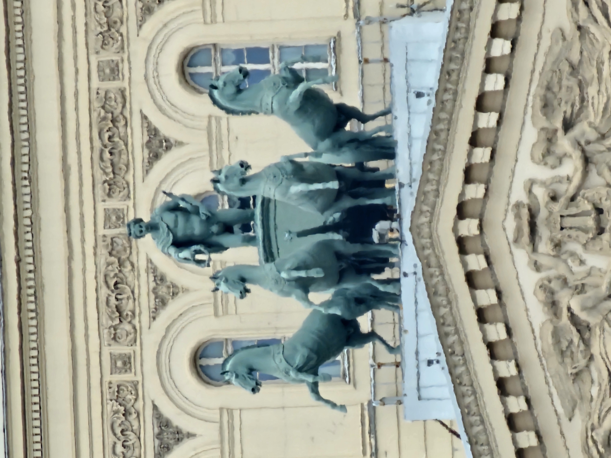 The image shows a statue of a man driving a four-horse chariot in Moscow, Russia. The statue is located on top of a building and is surrounded by ornate architectural details. The sky is visible in the background through the windows.