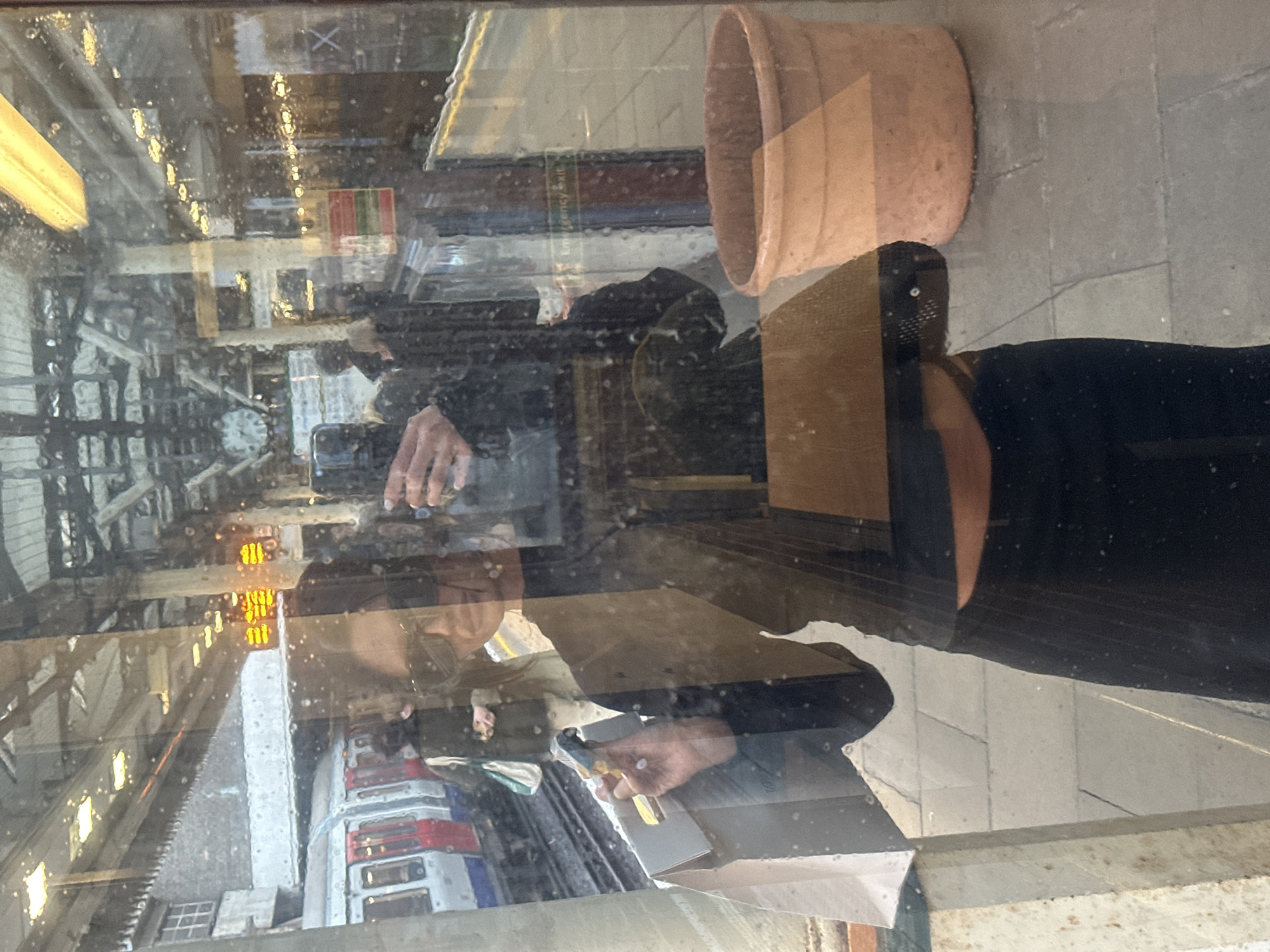 The scene depicts a London Underground station platform in the City of London, United Kingdom, viewed through a glass panel covered in water droplets.
In the foreground, the reflection of an individual is prominent, actively taking a photograph with a dark smartphone held in their right hand. This person wears dark sunglasses, a dark long-sleeved top that exposes the midriff, and dark trousers or a skirt. Their left hand holds a light-colored paper shopping bag with a cut-out handle.
Behind the reflection, on the platform, a London Underground train with characteristic red and white livery is visible to the left, stopped at the tracks. Several indistinct figures are present on the platform, one near the train and others further down. The station features overhead metal girder structures, artificial lighting fixtures, and a partially visible large, circular station clock with Roman numerals. Digital display boards exhibiting orange text are discernible in the distance. To the right of the reflection, a large terracotta plant pot rests on the grey platform surface, and a white "Emergency exit" sign is visible on a glass partition or door.