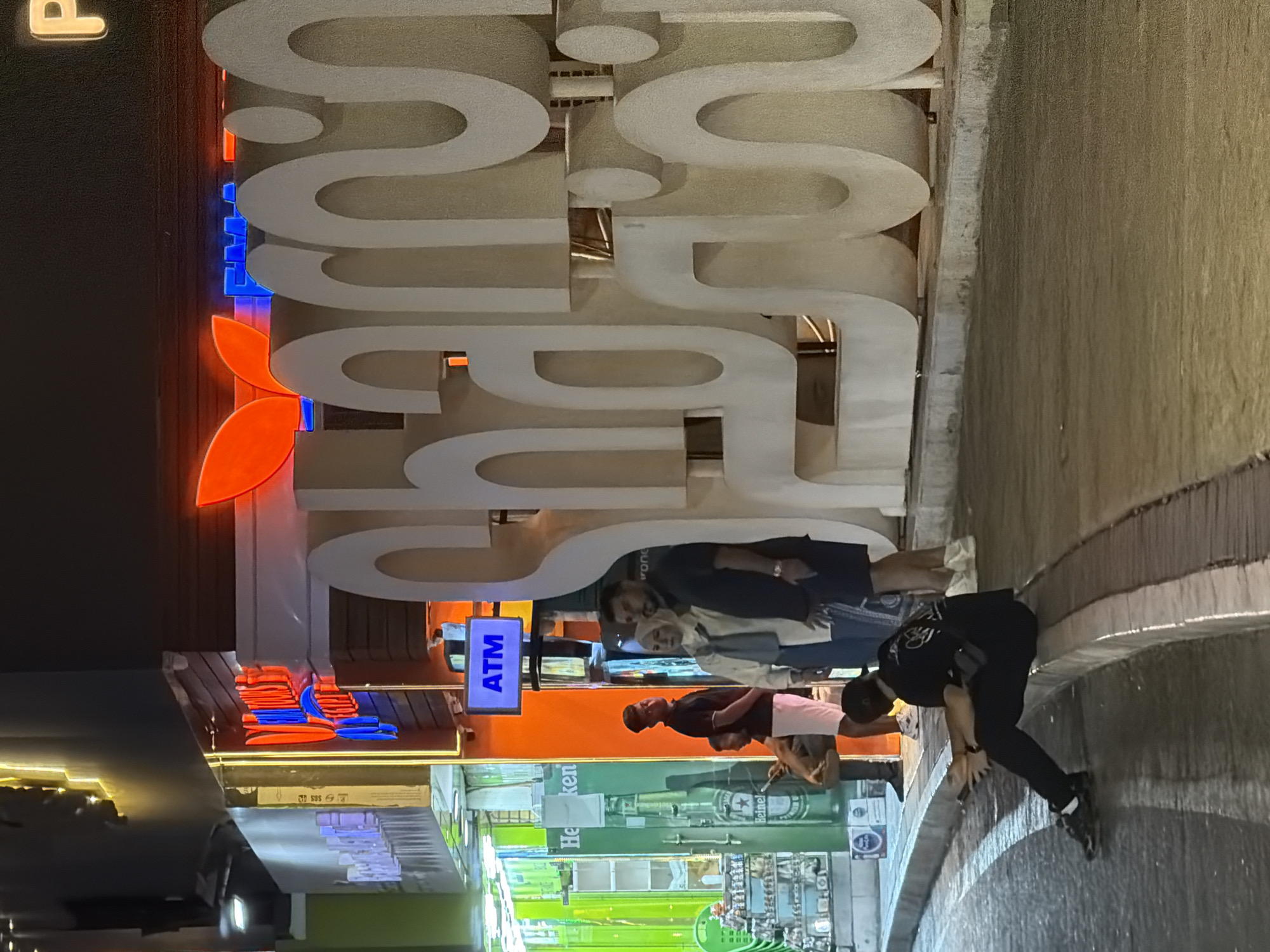 The image depicts a nighttime scene in Sharm El Sheikh, Egypt, focusing on a large, illuminated sign that spells out "Sharm" in Arabic and English. The sign is white and three-dimensional, with some letters being significantly larger than others. In the background, a building with red and blue neon lights is visible, possibly indicating shops or businesses. A blue "ATM" sign is also present.
In the foreground, several people are gathered. A couple stands near the large sign, a man in a dark t-shirt and white shorts looking towards the viewer, and a woman in a hijab and a light-colored outfit beside him. To their left, a man in a black t-shirt and white shorts is leaning against a green, illuminated display that appears to be advertising Heineken. Further to the left, behind a counter, are shelves stocked with various items, suggesting a store.
On the right side of the image, a person is seated on the curb, facing away from the viewer, looking at a mobile phone. This individual is wearing black clothing and appears to be relaxed. The ground is a paved street with distinct curbing. The overall atmosphere is lively, with bright artificial lighting illuminating the area.