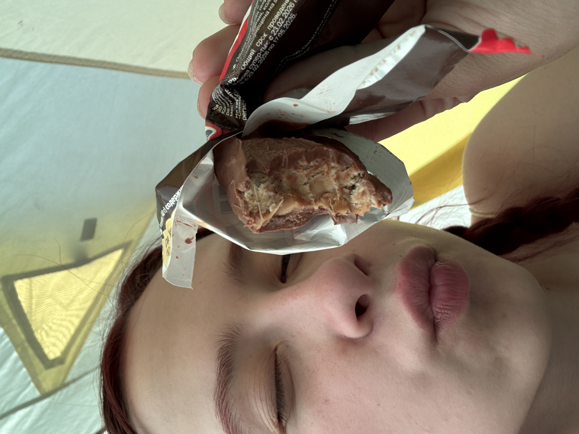 Here is a description of the image:
A close-up shot focuses on a person with fair skin, closed eyes, and red-tinted brown hair. They are holding a partially unwrapped chocolate bar in front of their face. The chocolate bar has a bite taken out of it, revealing a nougat-like filling. The packaging appears to be of a Mars-brand candy bar, with some Russian text visible on it. The background features what seems to be the inside of a tent, with light filtering through its fabric. Based on the OCR text, one can infer that this candy bar was made in the Khvalynsk, Russia area.