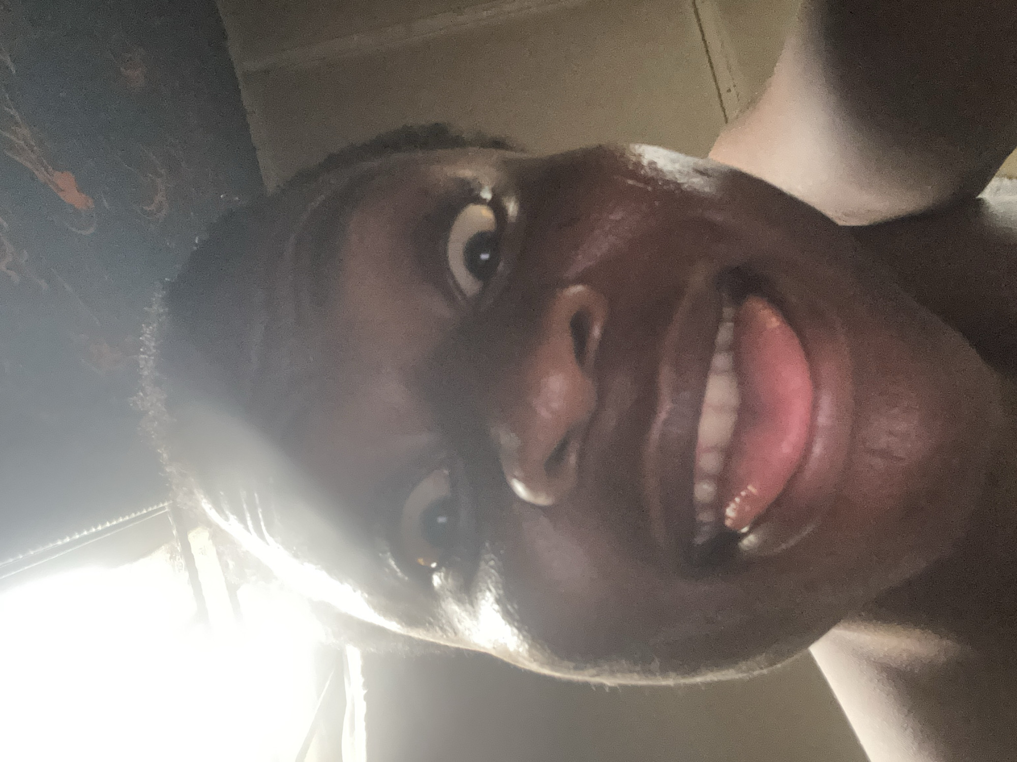 A close-up, rotated portrait depicts a person with dark skin, captured from a low angle looking upwards. The individual's head is tilted significantly, with their face filling the majority of the frame. They are making an exaggerated facial expression, characterized by widely opened eyes; the eye on the left side of the image appears particularly wide. The mouth is open, revealing white teeth and a pink tongue, which is either partially extended or resting on the lower lip.
A strong, bright light source originates from the lower left portion of the image, casting prominent highlights on the person's forehead, nose, and cheeks, and creating clear reflections in their eyes. The background is dark, out of focus, and largely indistinct, suggesting an indoor setting with uneven lighting. Hints of textured, dark surfaces, possibly a wall or ceiling, are visible in the upper left background. No specific clothing or additional objects are discernible. The overall mood conveyed is one of playfulness, surprise, or silliness due to the expressive features. There is no visible text.