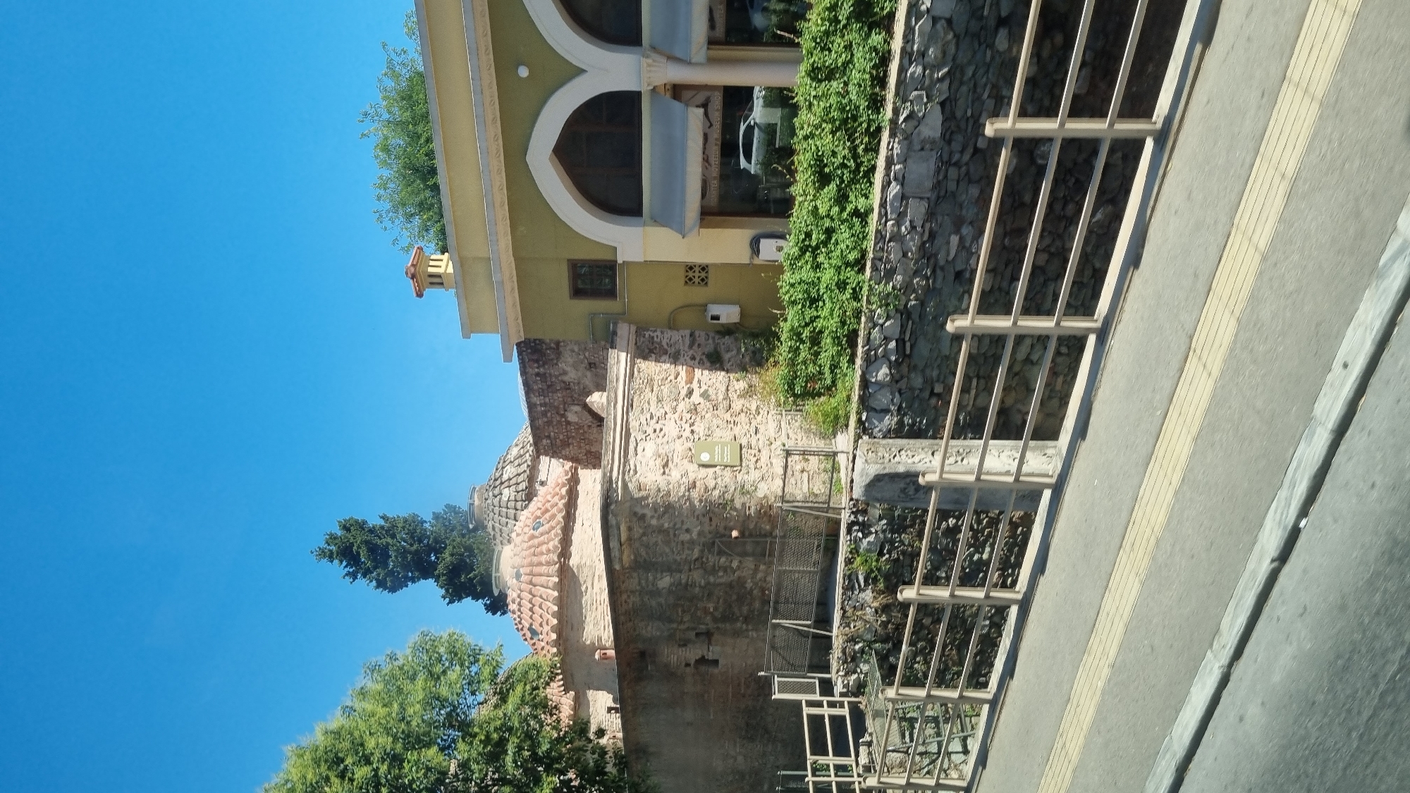 This is a medium shot, taken from a slightly elevated angle, of a historic building complex in Thessaloniki, Greece, on a clear, sunny day. The foreground shows a paved road with a textured yellow tactile paving strip and a concrete curb. A metal railing with horizontal bars separates the road from a stone retaining wall and a small, dry creek bed or ditch.
Behind the retaining wall are several structures. To the left, a large stone wall forms the base of buildings with red-tiled, domed roofs, characteristic of traditional architecture. A tall, slender evergreen tree stands behind these domes. To the right, a lighter-colored building with yellow facade and arched windows and doorways is visible. This building appears to be a cafe or shop, with awnings over the windows and a reflection of a white car in the glass. Lush green bushes and plants grow in front of this building and along the top of the stone wall.
The sky is a vibrant, clear blue. The overall impression is a well-preserved historical site integrated into a modern urban environment, likely in the Thessaloniki Municipal Unit. No people are visible in the frame.