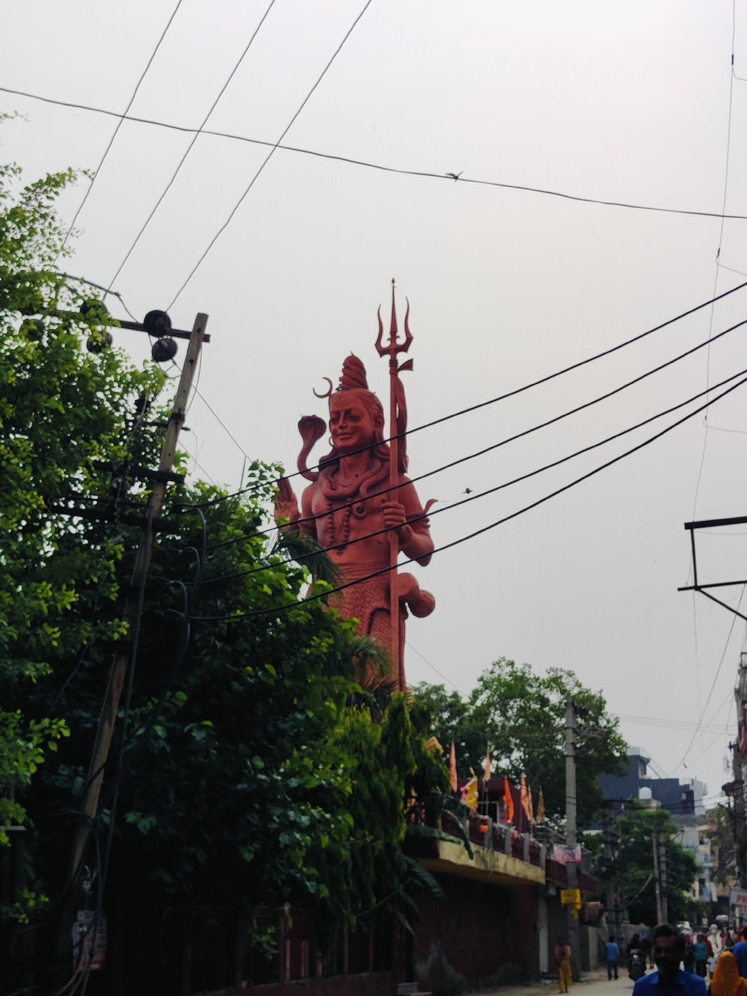 The image shows a large reddish statue of the Hindu deity Shiva, holding a trident, towering over a street scene. The statue is the central focus, with its imposing presence dominating the upper right portion of the frame. In the foreground and to the left, lush green trees partially obscure a wooden utility pole with electrical wires strung across the sky. The wires create a crisscrossing pattern against the overcast sky. Below the statue, a building with a balcony adorned with flags can be seen, suggesting a religious or cultural site. Further down the street, several people are visible, some walking and others on motorbikes, indicating a typical urban environment. The overall impression is of a spiritual landmark integrated into the daily life of a bustling city street.