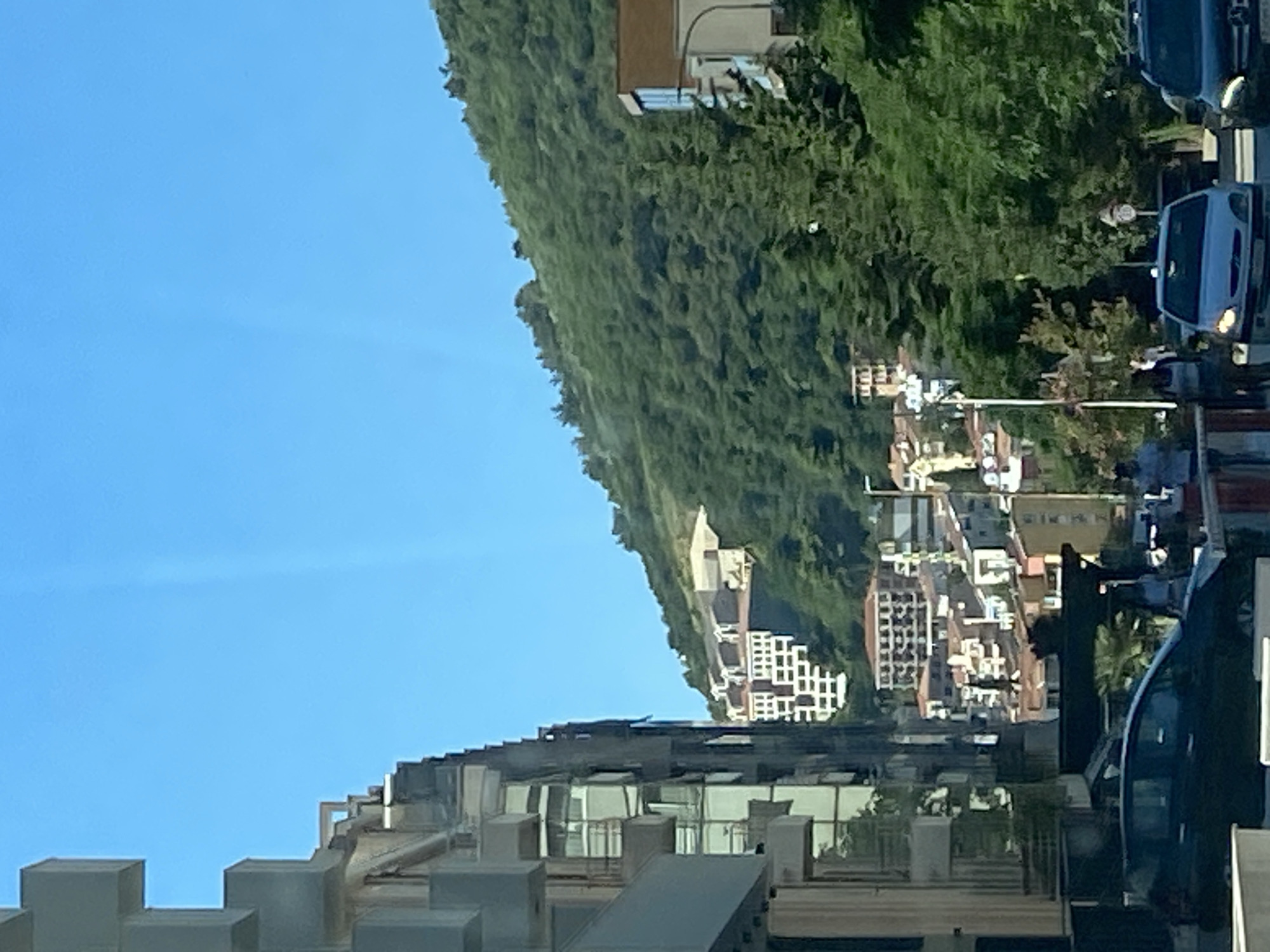 The image shows a street scene in Budva, Montenegro, on a clear, sunny day. A modern, light-colored apartment building with multiple balconies dominates the left side of the frame. In the background, a lush green hillside rises, dotted with buildings and trees. The buildings on the hillside appear to be a mix of residential and hotel structures, with white and terracotta-colored facades. A dark-colored car is parked on the street in the foreground, and a light-colored car and a dark SUV are visible driving on the road. Several people are present on the sidewalk, some walking and some standing. The overall impression is of a vibrant Mediterranean coastal city with a mountainous backdrop.