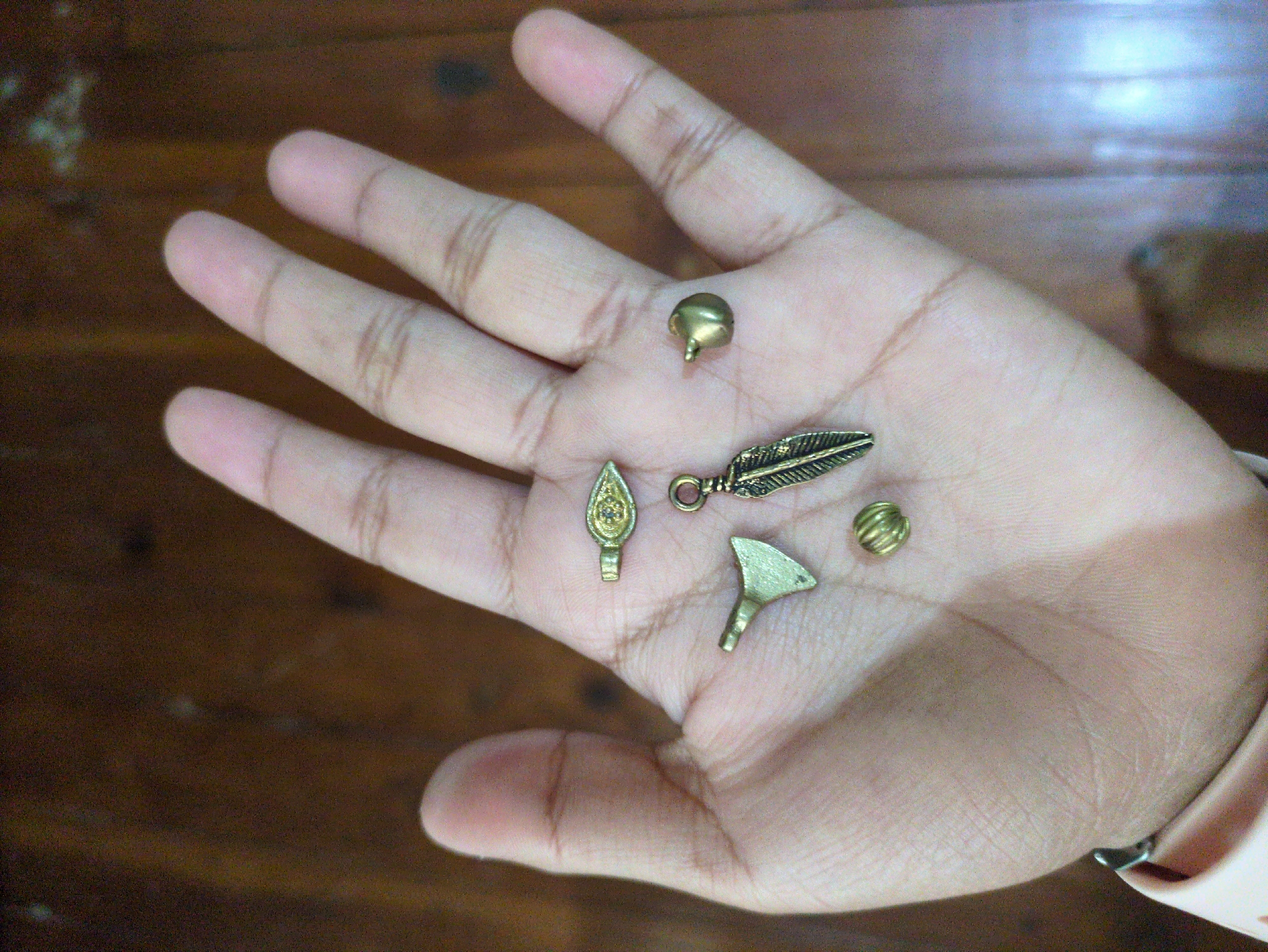 A human hand, palm up, displays a collection of small, antique-looking metal charms. These charms include a miniature axe, a round bead with ribbed texture, a feather pendant, a bell-shaped charm, and a decorative, elongated oval charm with intricate etchings. The hand is resting on a wooden surface, possibly a table or floor, with a visible wood grain pattern. The lighting is dim, casting soft shadows. The image appears to be a close-up shot, focusing on the palm and the charms. There is no information available to determine the specific location of Haini, India.