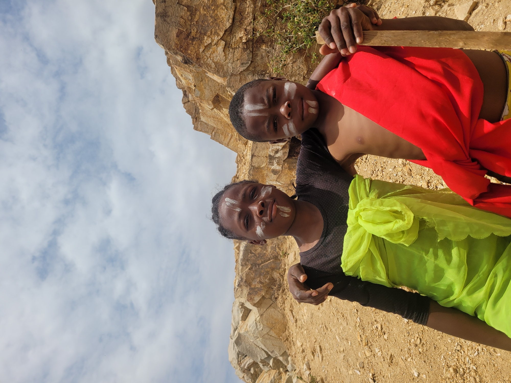 Two children stand in front of a rocky cliff face under a partly cloudy sky. Both children have white face paint markings on their foreheads and cheeks. The child at the top of the image is shirtless and draped with a red cloth. They are holding a wooden object. The child at the bottom of the image wears a black shirt and is draped with a green cloth. The ground is sandy and rocky. The location is Barakin Akawo, Nigeria.