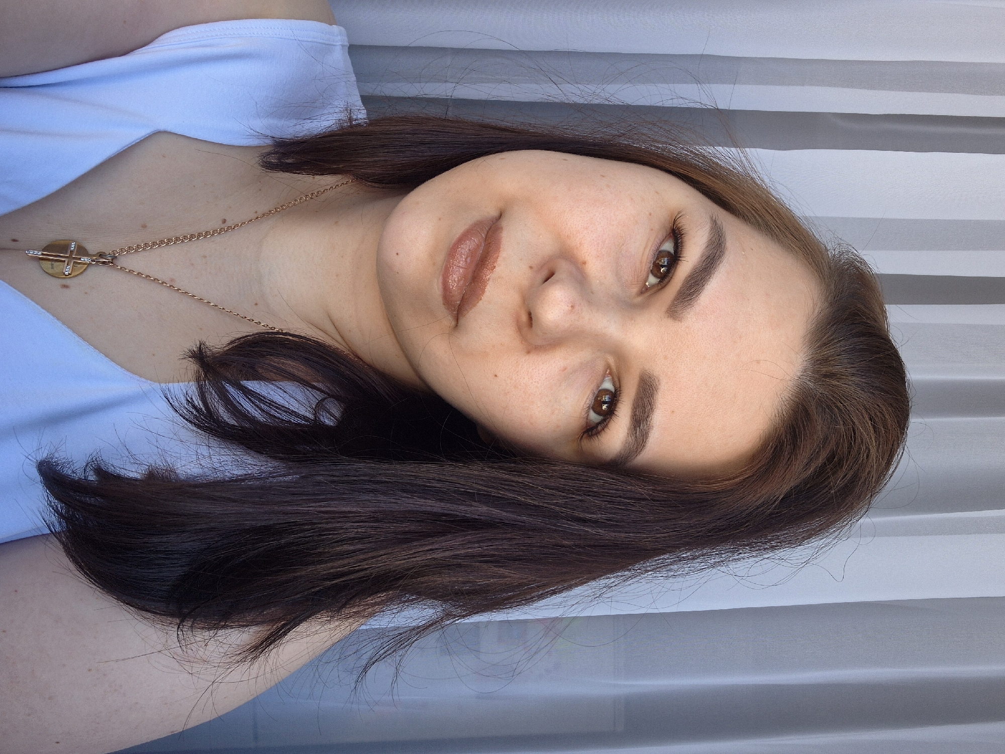 A woman with shoulder-length, brown hair and light skin is featured in a close-up portrait. She is wearing a light blue top and a gold necklace with a round pendant bearing a cross design. The woman is angled slightly, her brown eyes gazing towards the viewer. The background consists of vertical, pale gray and white stripes, possibly a window covering. The location is not discernible from the image. There are no indications of Tobolsk, Russia.