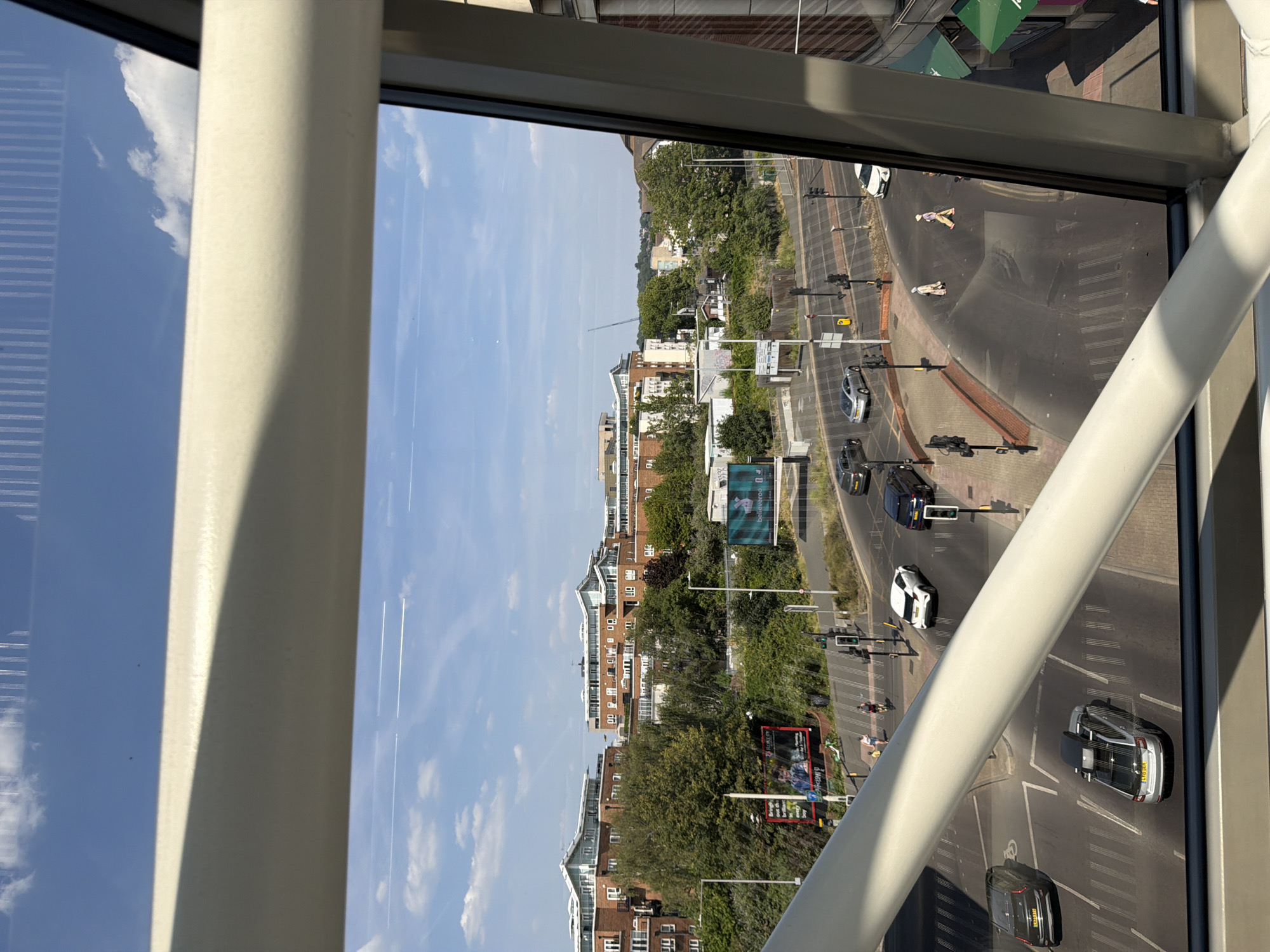 This is a daytime, high-angle shot taken from inside a modern, elevated walkway or building, looking out over a city street scene. The foreground is dominated by a thick, cream-colored horizontal beam that bisects the frame, casting a shadow. Through the glass of the walkway, we see a multi-lane road with several cars in motion, including a silver hatchback with a roof box, a dark sedan, a white SUV, and a silver sedan. Pedestrians are visible crossing the street at a marked crosswalk, and traffic lights are positioned at the intersection.
Beyond the road, there are more vehicles and infrastructure, including traffic signs and streetlights. In the mid-ground, lush green trees line the road, interspersed with brick apartment buildings featuring modern architecture and white-tiled roofs. One large billboard features an advertisement with two men. In the background, a construction crane is visible, suggesting ongoing development in the area. The sky is blue with scattered white clouds and contrails from airplanes. The overall impression is a contemporary urban environment on a sunny day. This appears to be a view of the United Kingdom, likely a city with modern developments.
