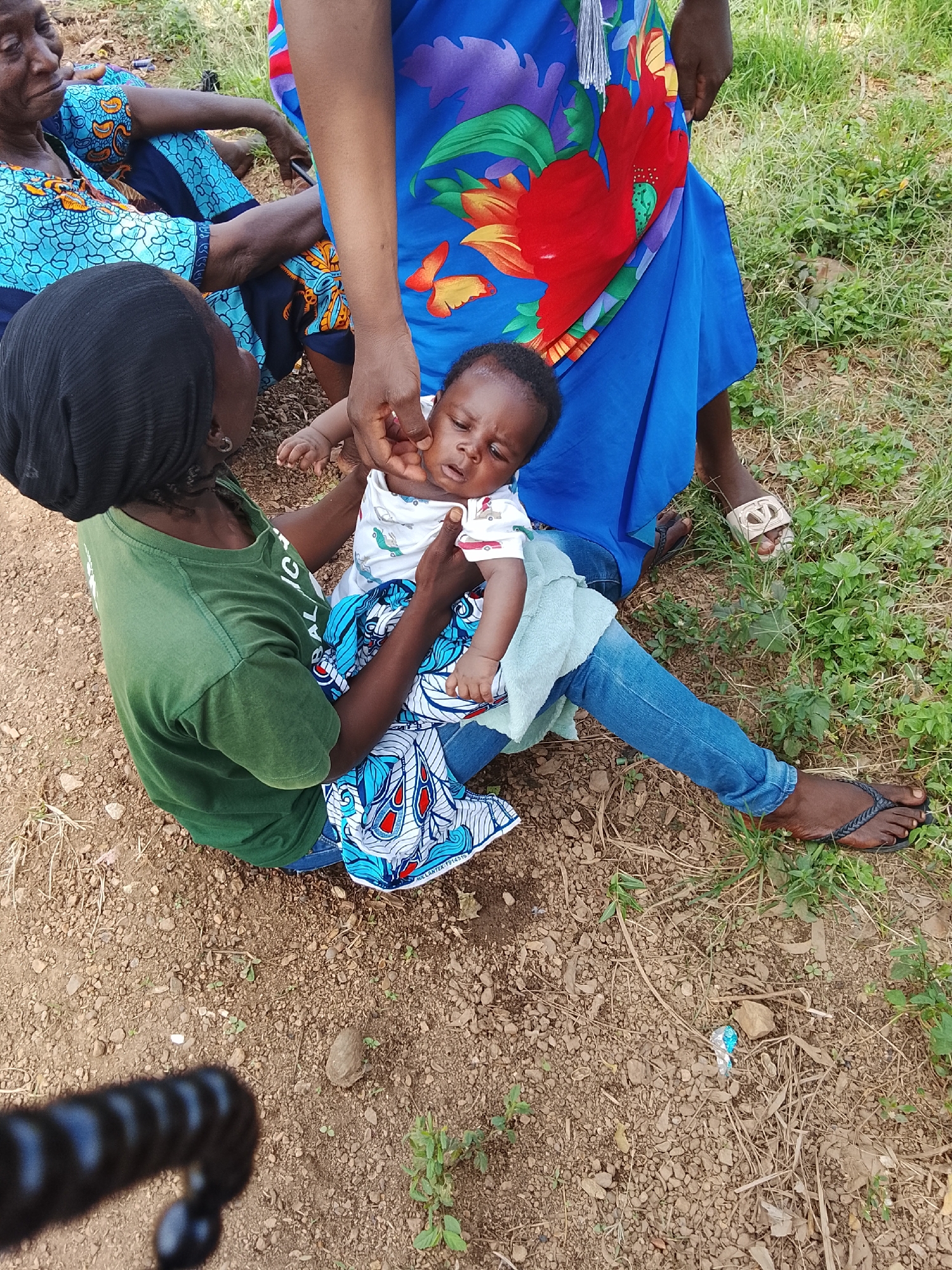This image shows a group of people gathered outdoors. The main focus is on a woman in a green t-shirt, holding a baby. The baby is wearing a white onesie with a subtle pattern and appears to be asleep or very relaxed. The woman holding the baby is wearing a black headscarf and blue jeans. Another woman, wearing a vibrant blue dress with a large floral print, stands behind them, with her legs and feet visible. She is wearing white sandals. To the left, partially visible, is another woman in a blue and orange patterned dress.
The setting appears to be a dirt and grass area, possibly a rural or semi-rural location. The ground is covered with loose soil, small stones, and patches of dry and green grass. There are some dry leaves and twigs scattered around. The lighting suggests it is daytime, likely outdoors with natural sunlight.
The activity appears to be a moment of care and rest. The woman is cradling the baby gently, and the presence of other people suggests a communal gathering. The overall mood seems calm.
There is no visible text in the image. The clothing of the people, particularly the women's dresses and the baby's onesie, features colorful patterns. One of the women's dresses has a blue and red floral design. The patterned fabric beneath the baby also has intricate blue, white, and red designs.